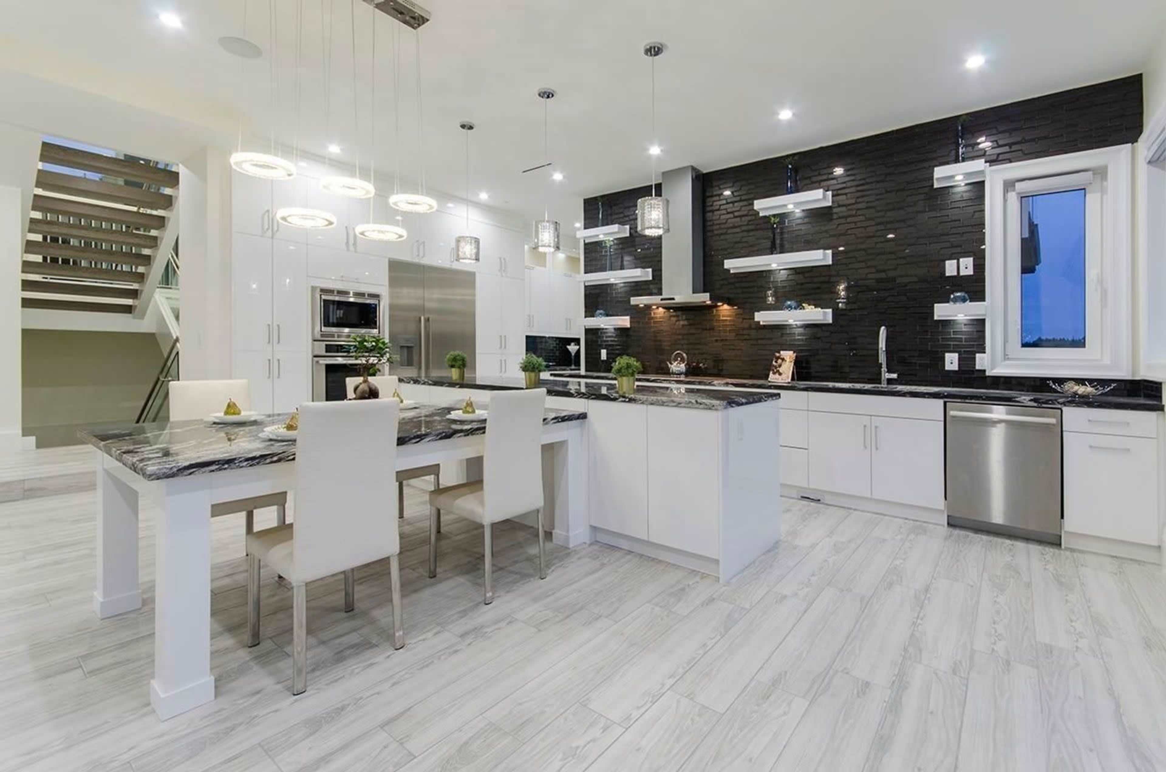 A modern kitchen featuring a large island with seating, sleek white cabinets, a black tiled wall, and contemporary lighting fixtures.