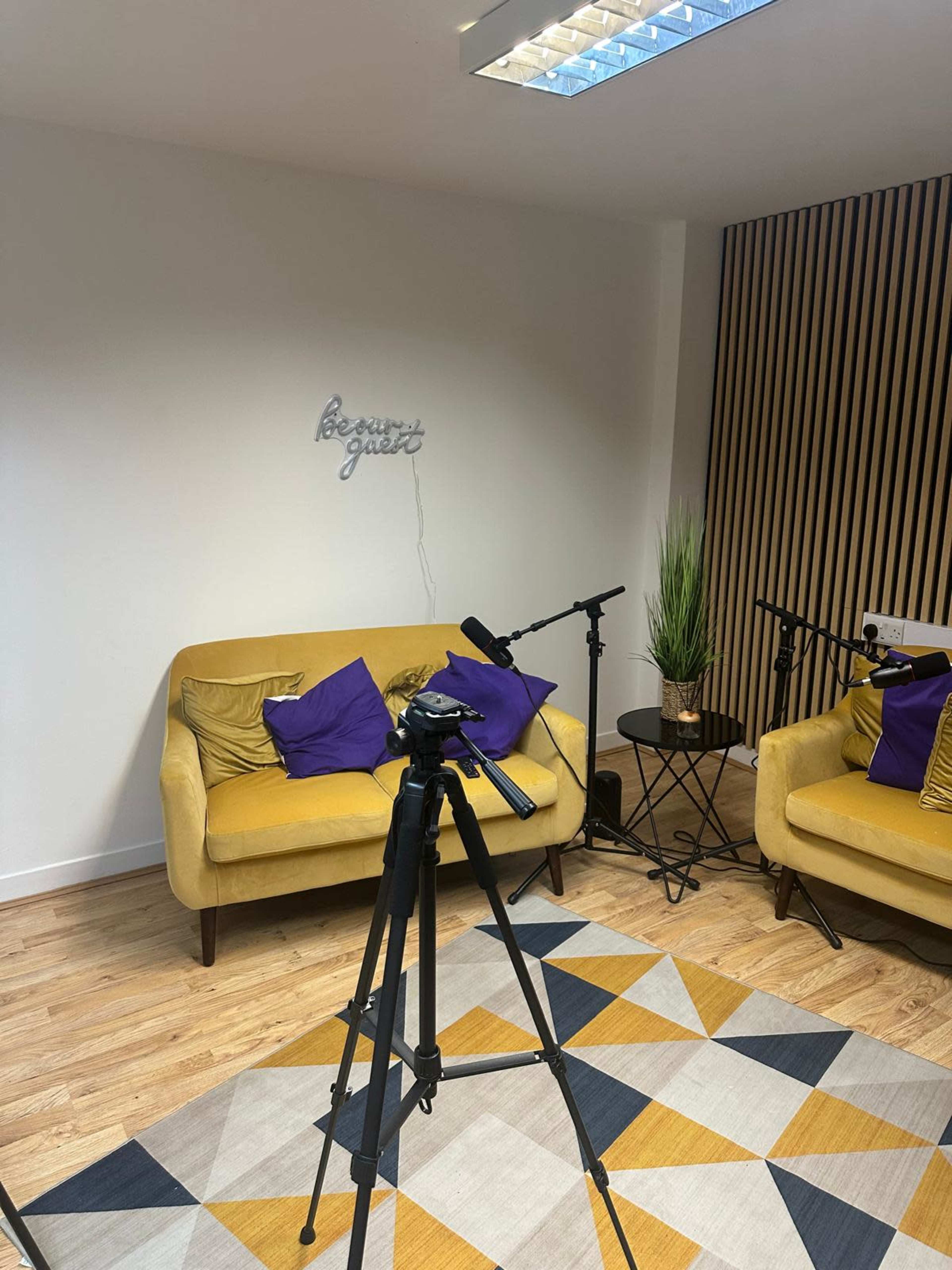 The image shows a small studio living area with two yellow sofas, several purple pillows, a tripod with a camera, and a patterned rug on the wooden floor.