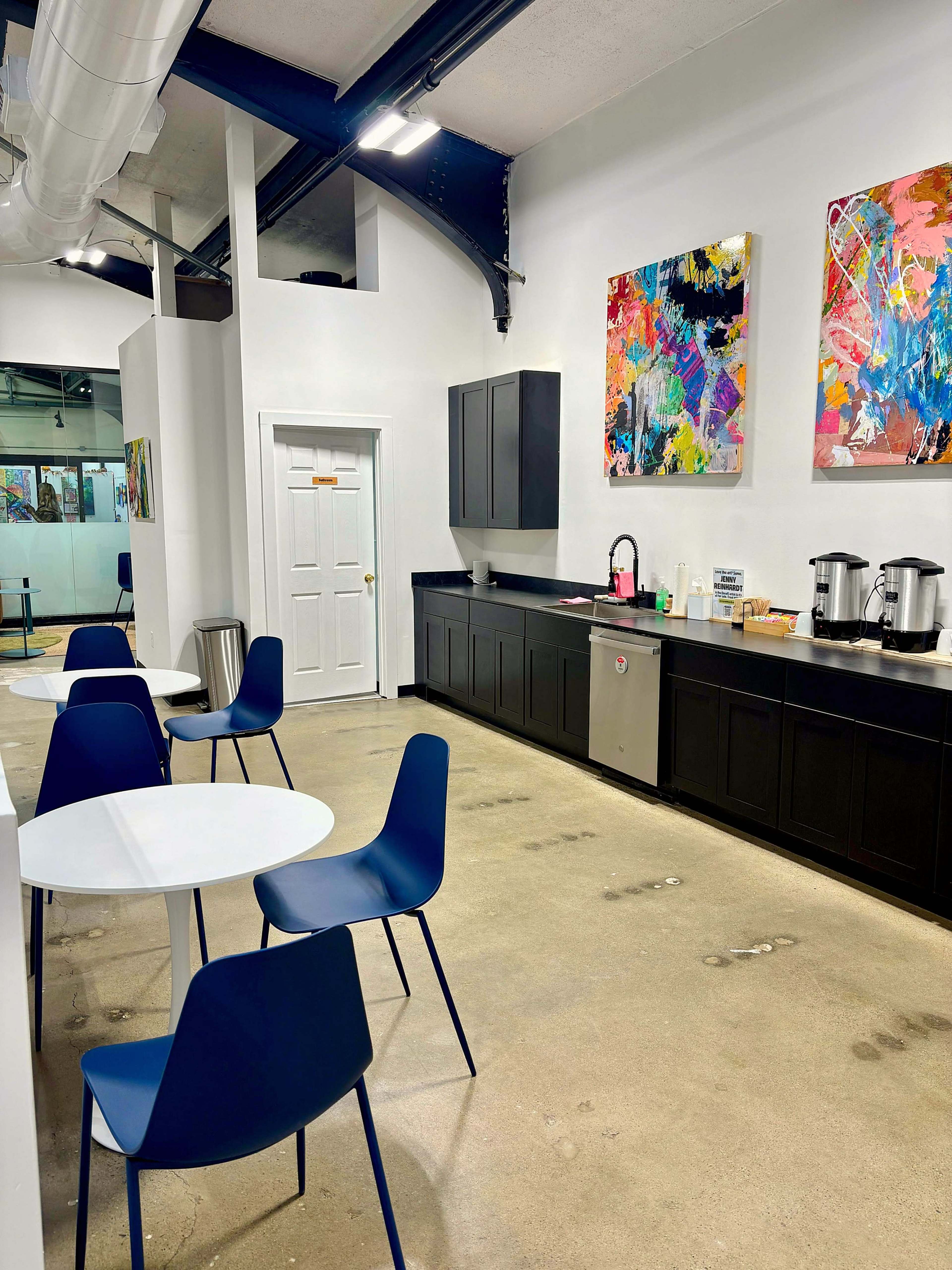 The image shows a modern kitchen or break area with black cabinetry, white walls, abstract paintings, and several round tables with blue chairs.