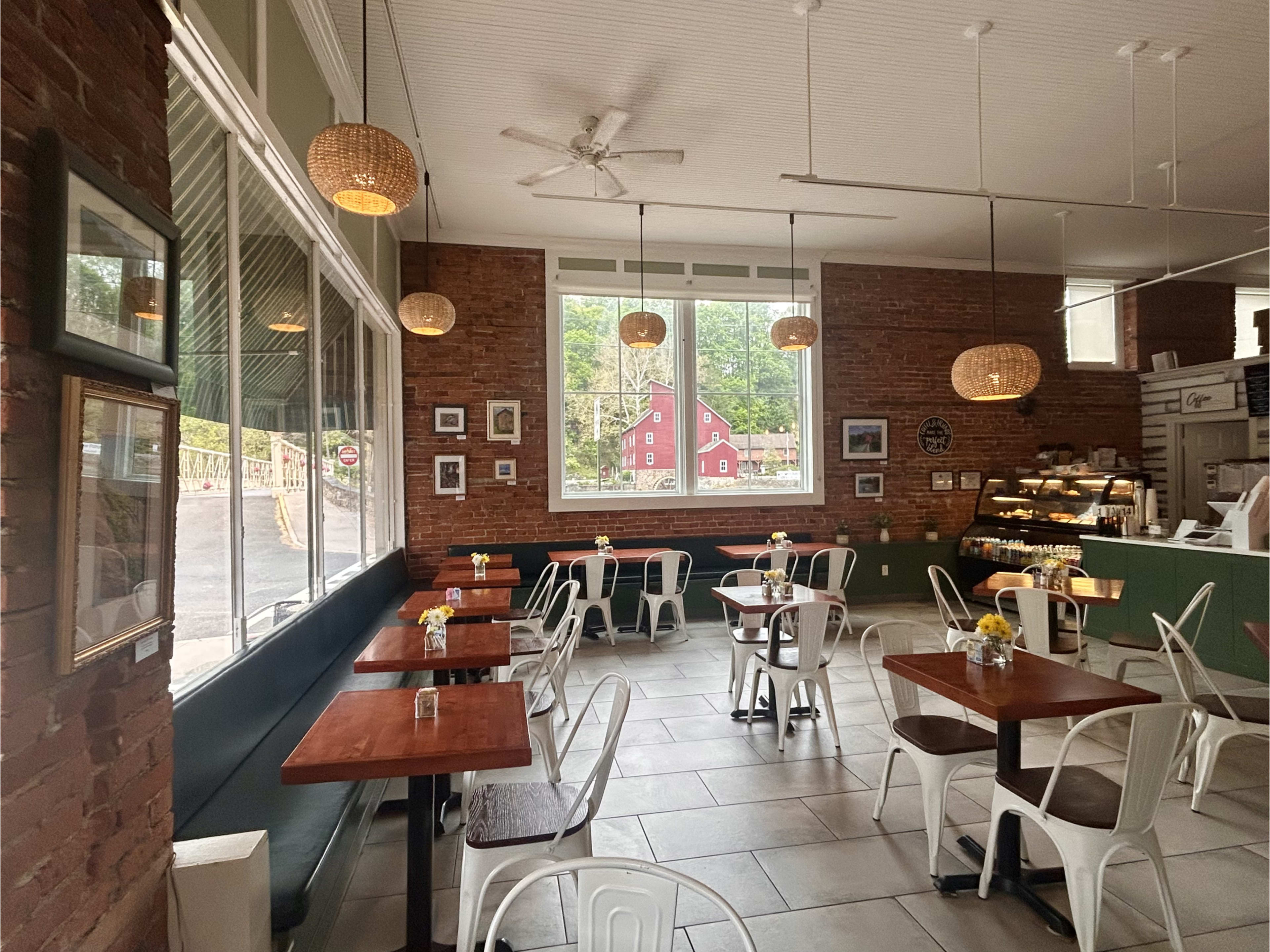 The image shows a cozy café with wooden tables, white chairs, and large windows displaying a view of a red barn outside.