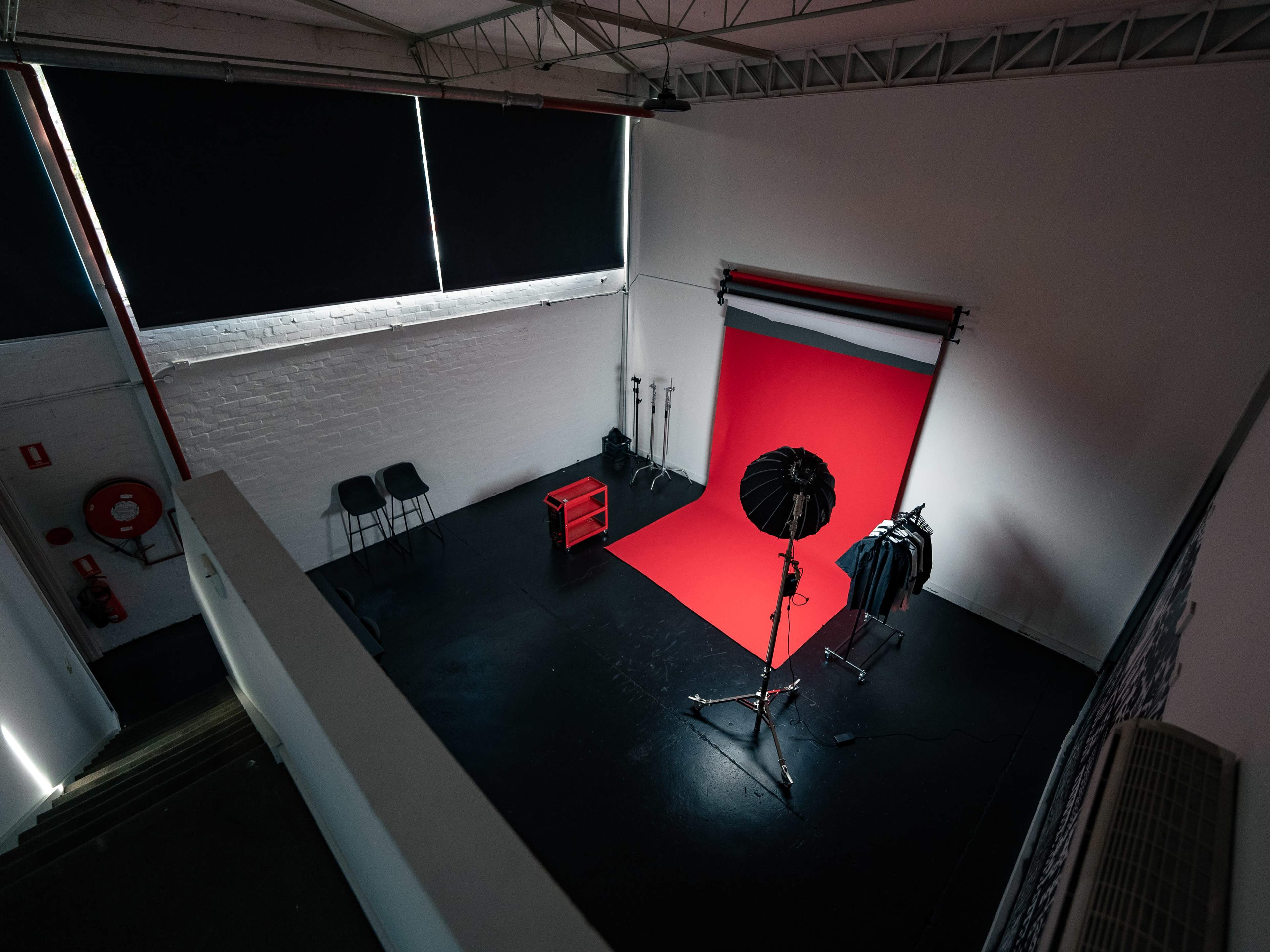An empty photography studio features a red backdrop, lighting equipment, and a few chairs arranged against a white wall.