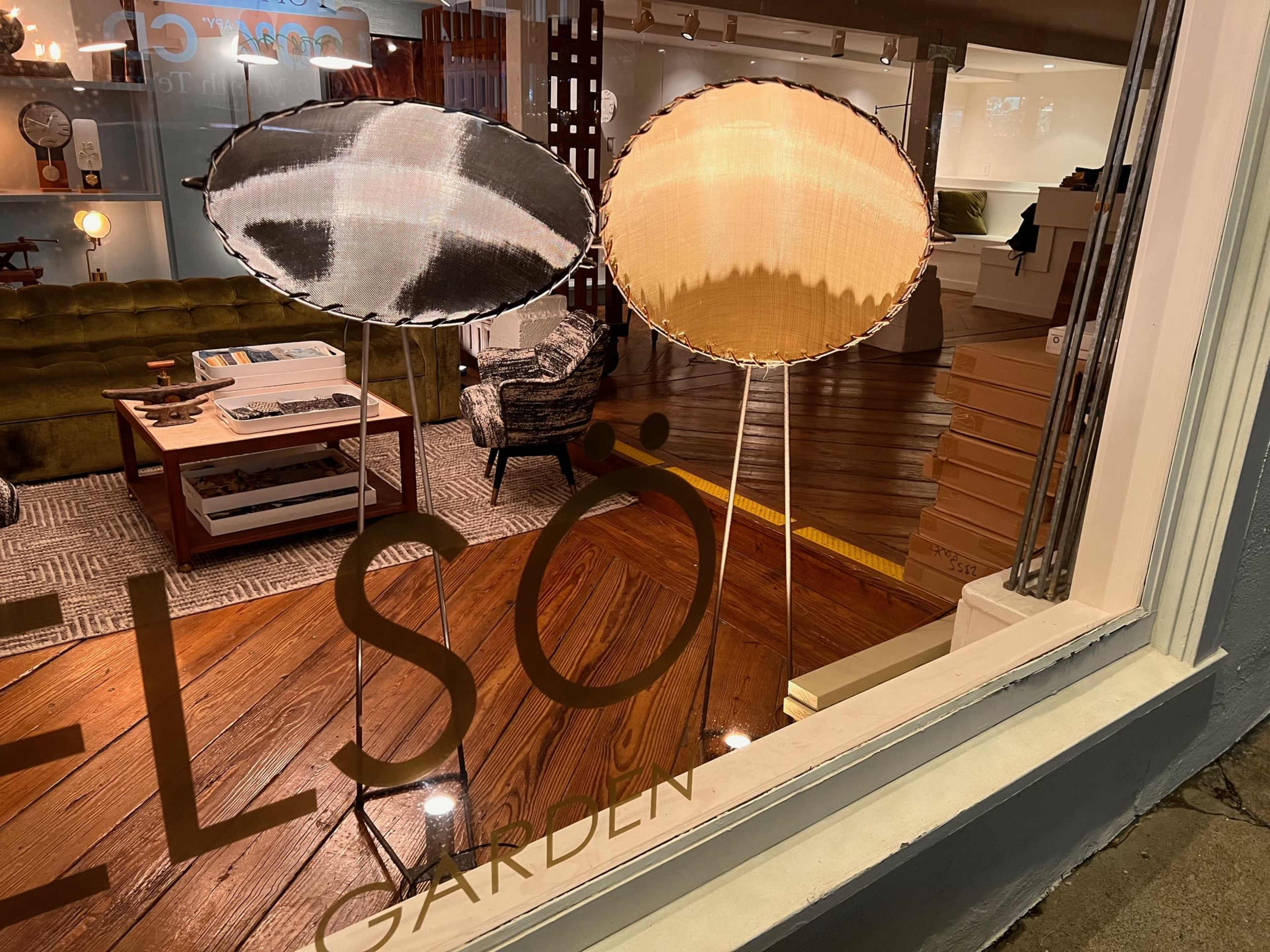 Two standing lamps with circular shades are displayed in a shop window, with wood flooring and furniture visible in the background.