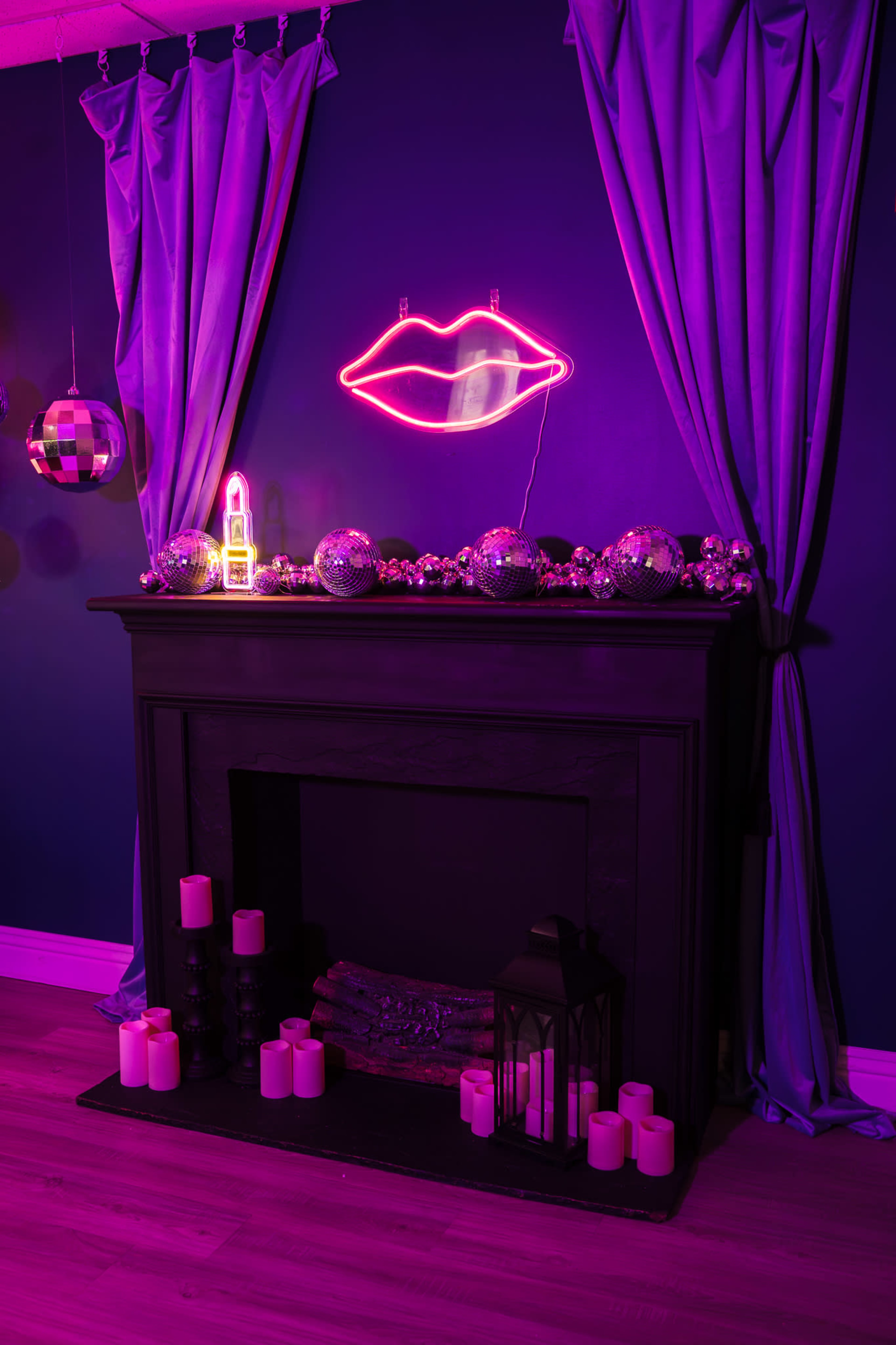 The image shows a fireplace decorated with candles, a lantern, and a string of disco balls, with a pink neon lips sign above it and curtains draped on either side.