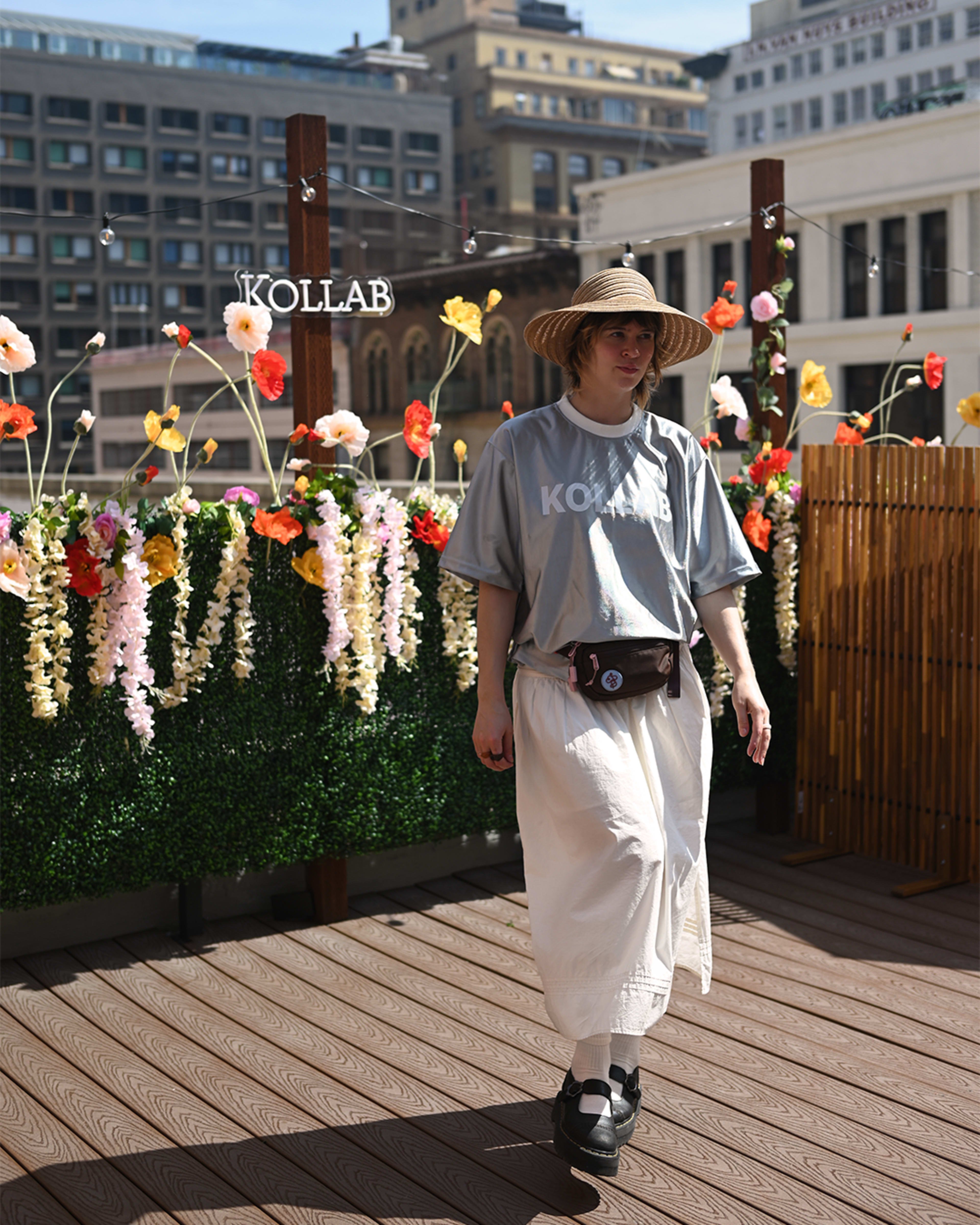 A person wearing a gray shirt and white skirt walks on a wooden platform decorated with colorful flowers and a sign that reads "KOLLAB."