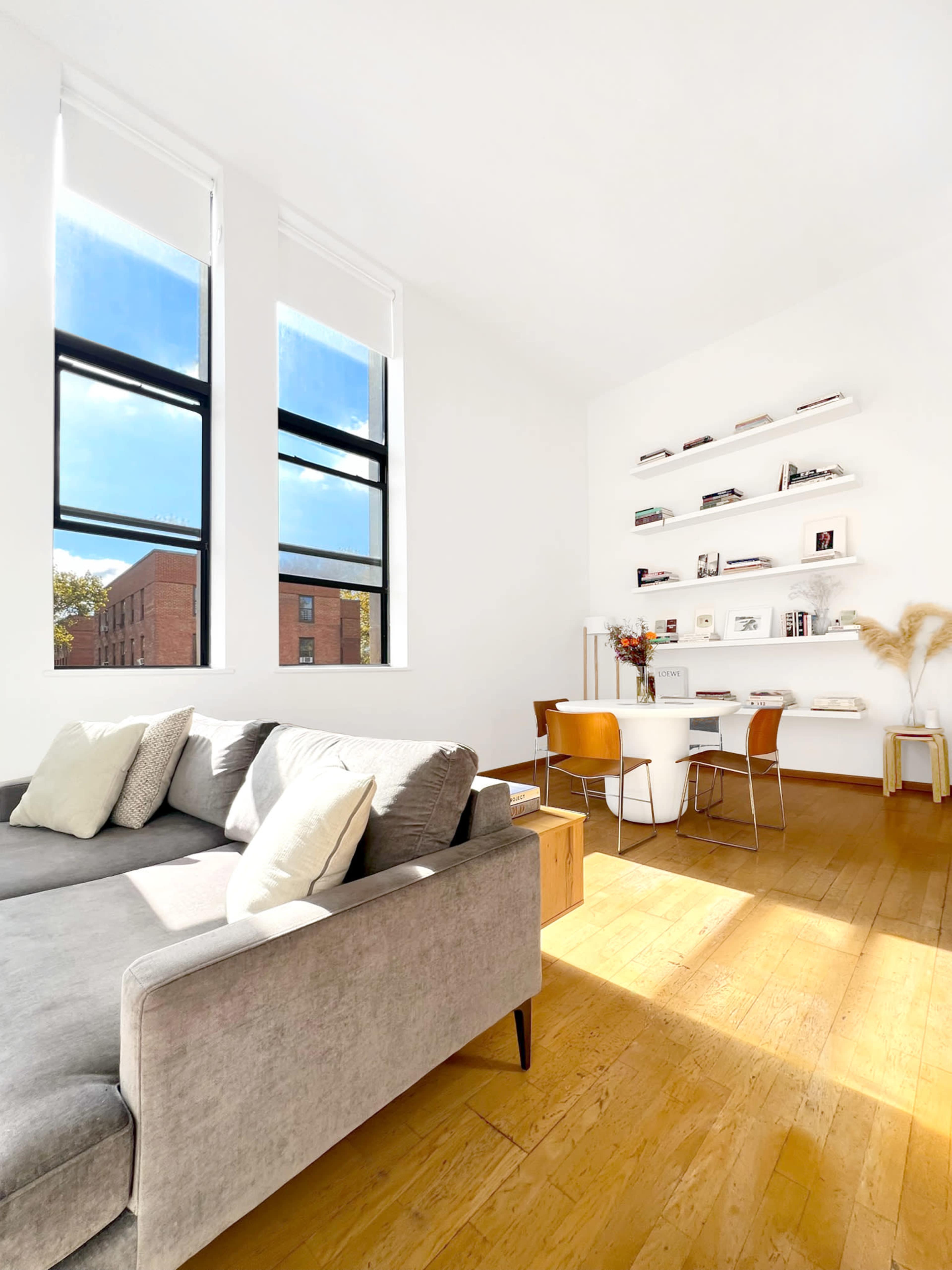 The image shows a bright, modern living space featuring a gray sofa, a small dining table with chairs, and shelves displaying books and decor against a white wall.