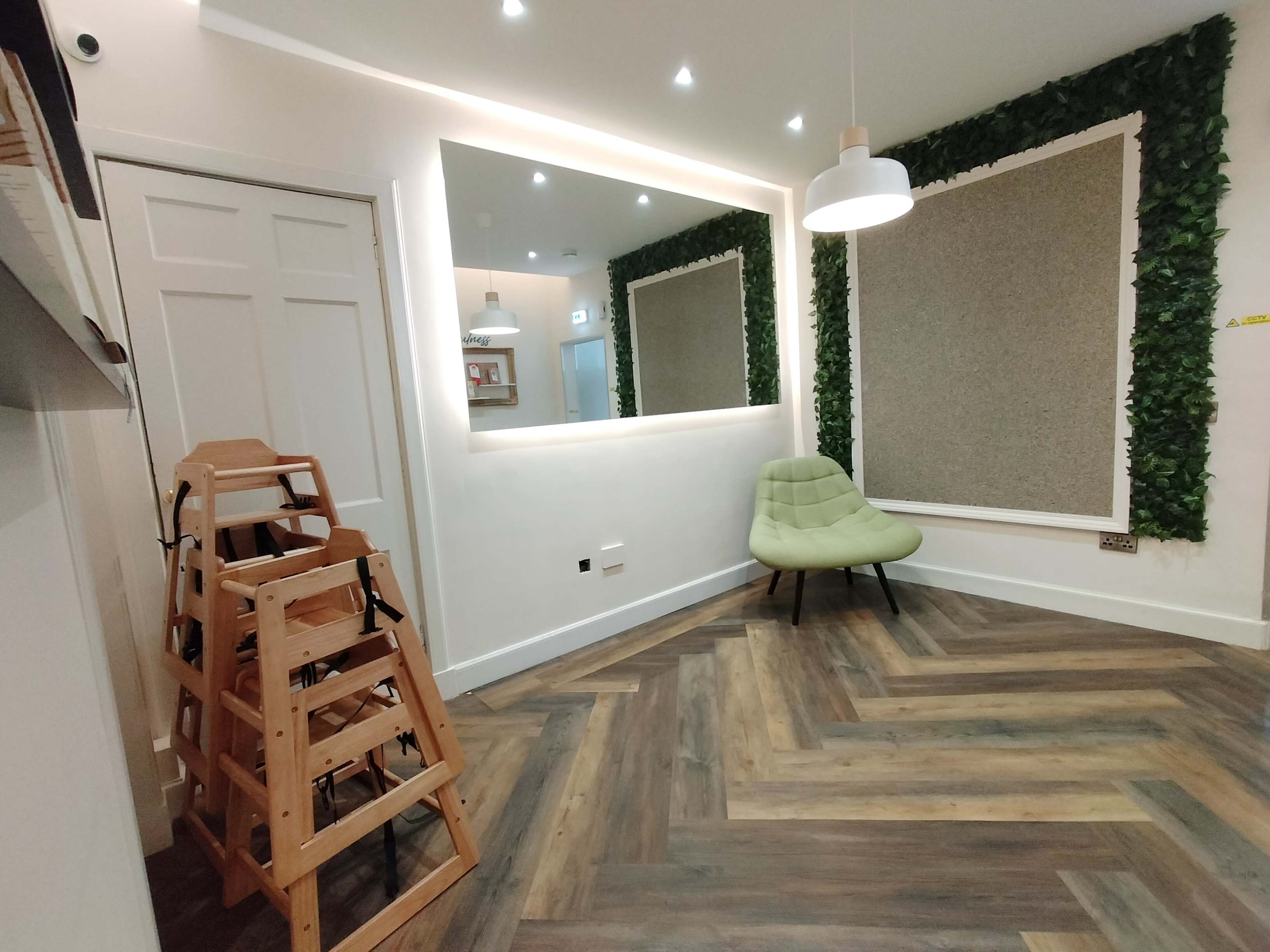 A modern room with a large mirror, green foliage on the walls, a single chair, and stacked high chairs in the corner, all set on patterned wooden flooring.