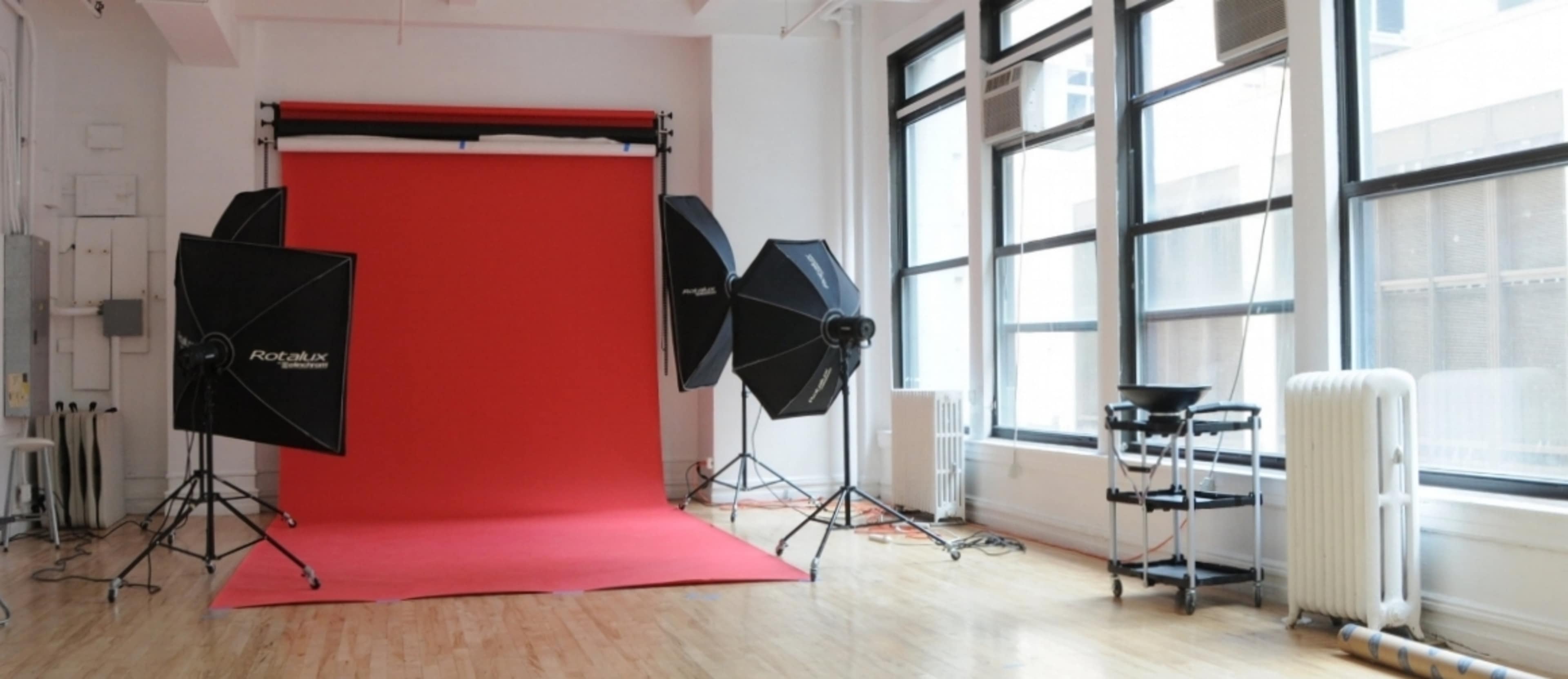 A photography studio with a red backdrop, various lighting equipment, and large windows allowing natural light to enter.
