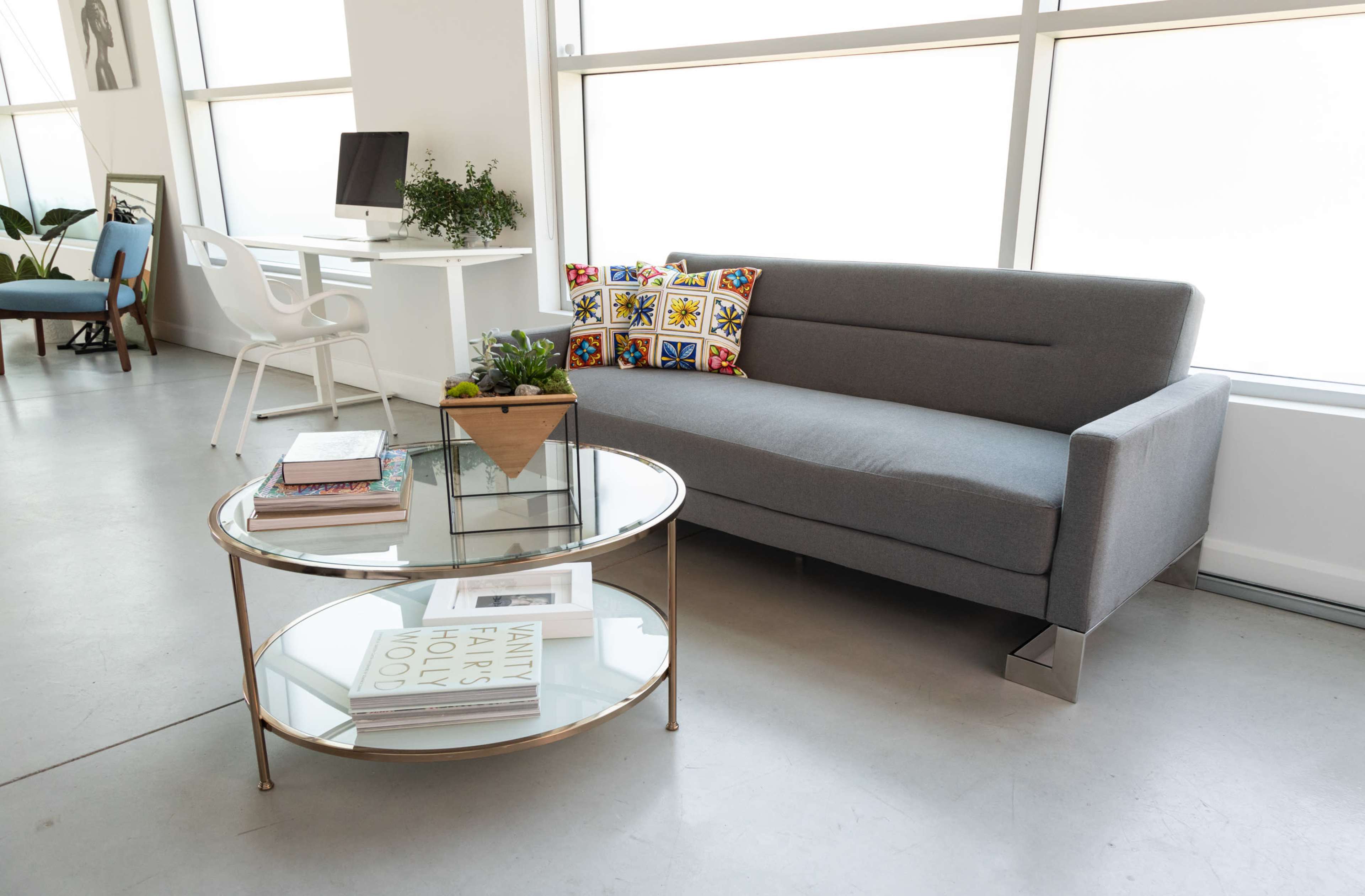 A modern living space featuring a gray sofa, a round glass coffee table with books and a plant, and a desk with a computer in the background.