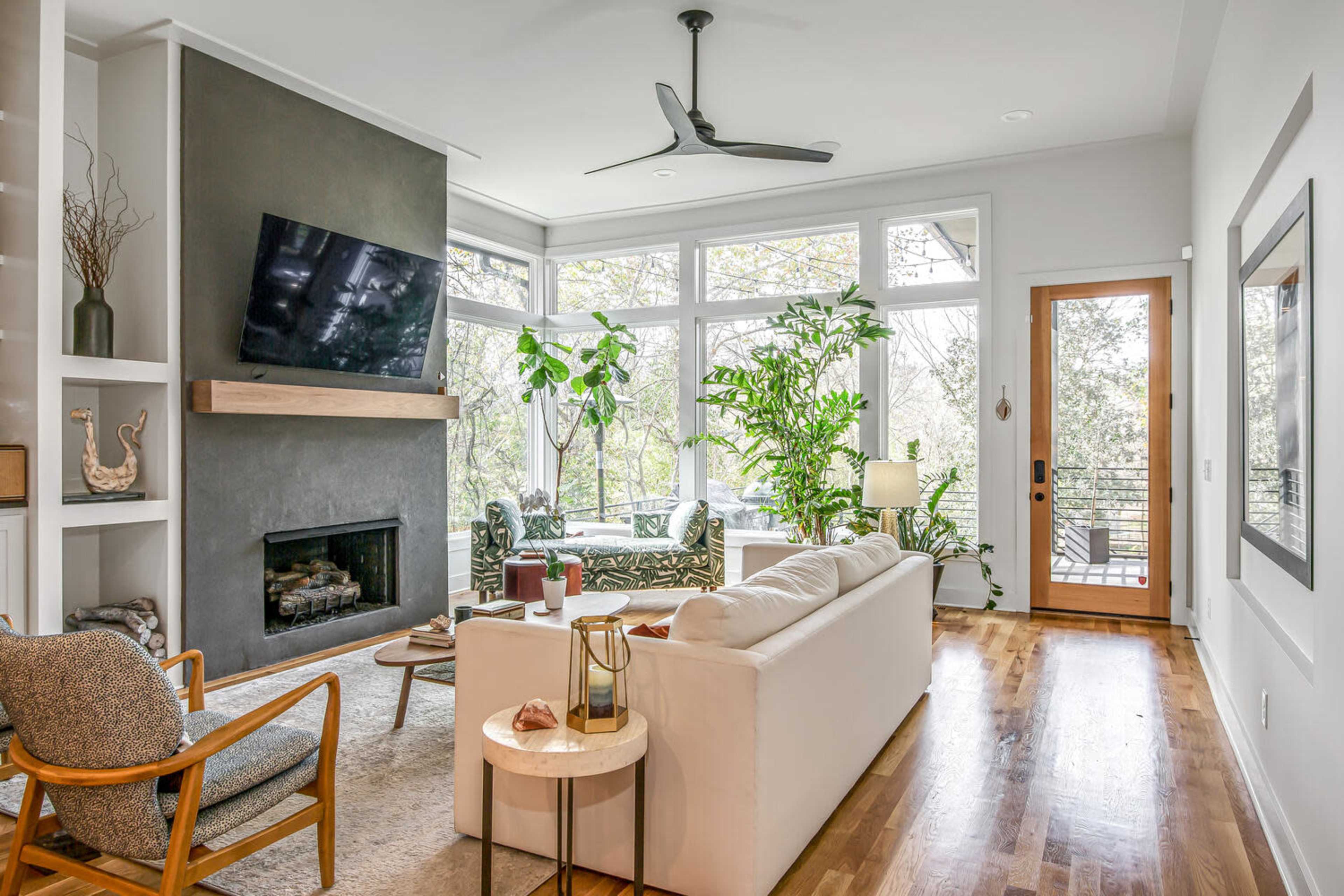 A bright living room features a large window, potted plants, a white sofa, and a wall-mounted TV above a fireplace.