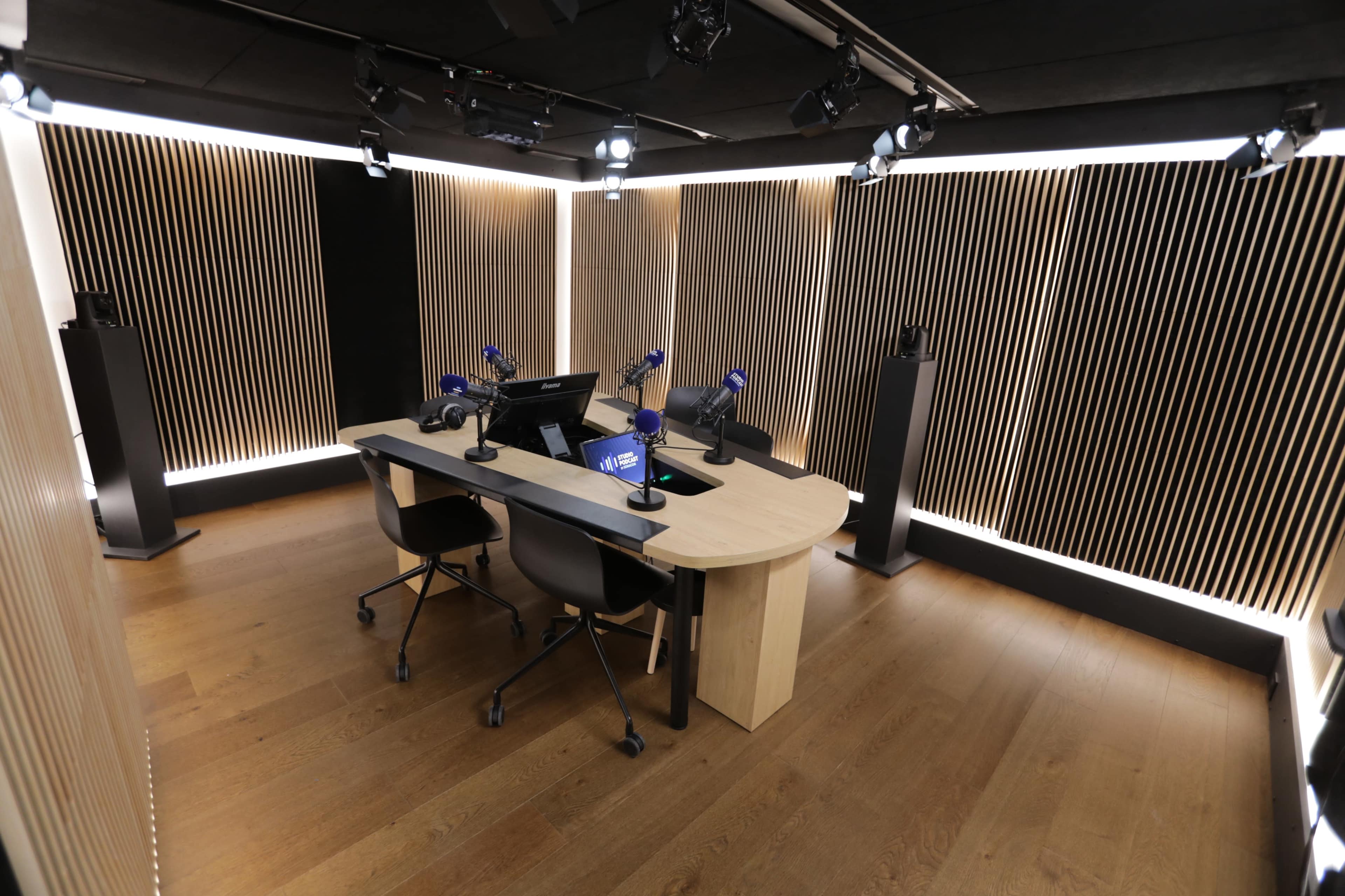 A modern radio studio with a curved desk, two chairs, and multiple microphones arranged in a sleek, wooden interior.