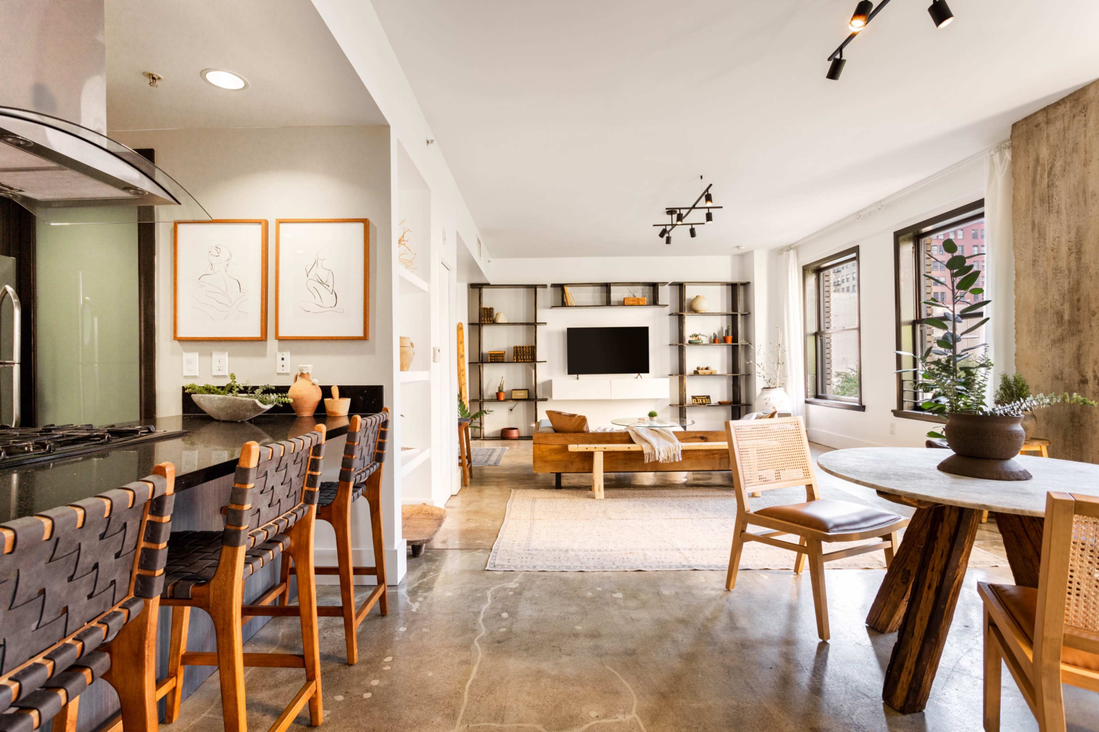 A modern living space with a kitchen bar, a dining table, and a seating area featuring a television and shelves against a wall of large windows.
