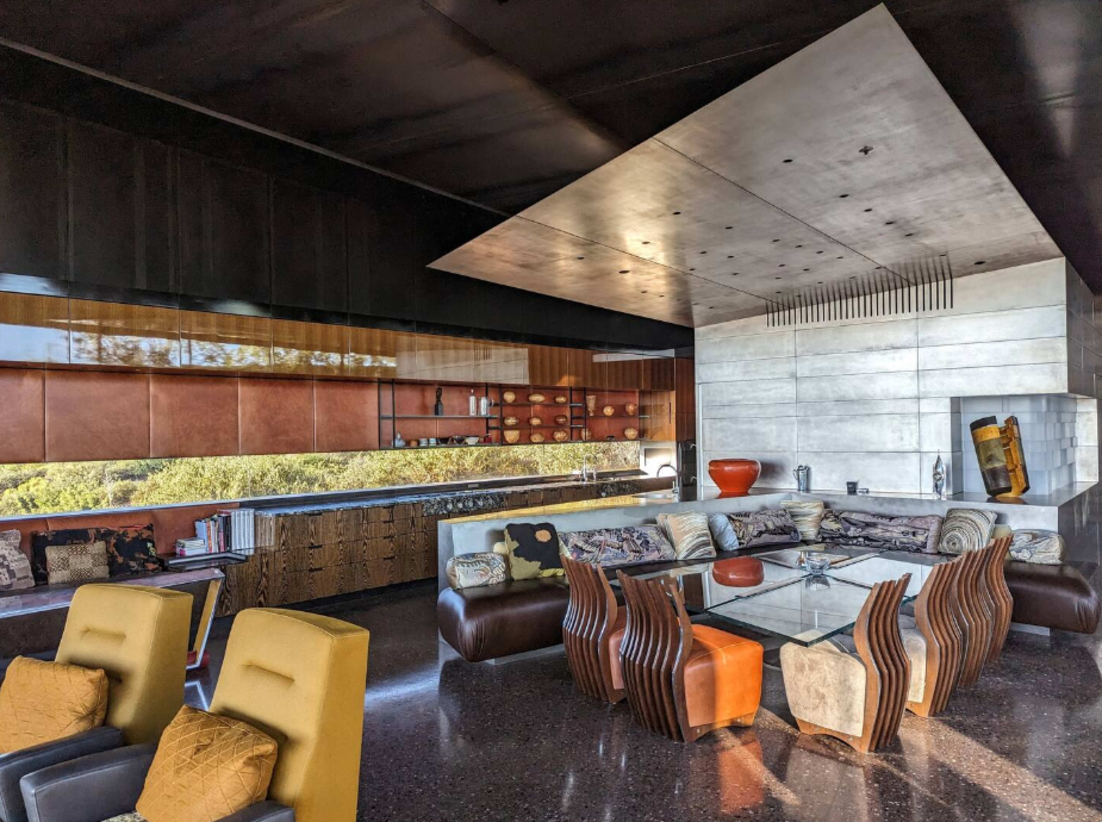 A modern living room with a glass table surrounded by stylish seating, featuring a sleek kitchen area with a contrast of materials and a view of greenery through large windows.