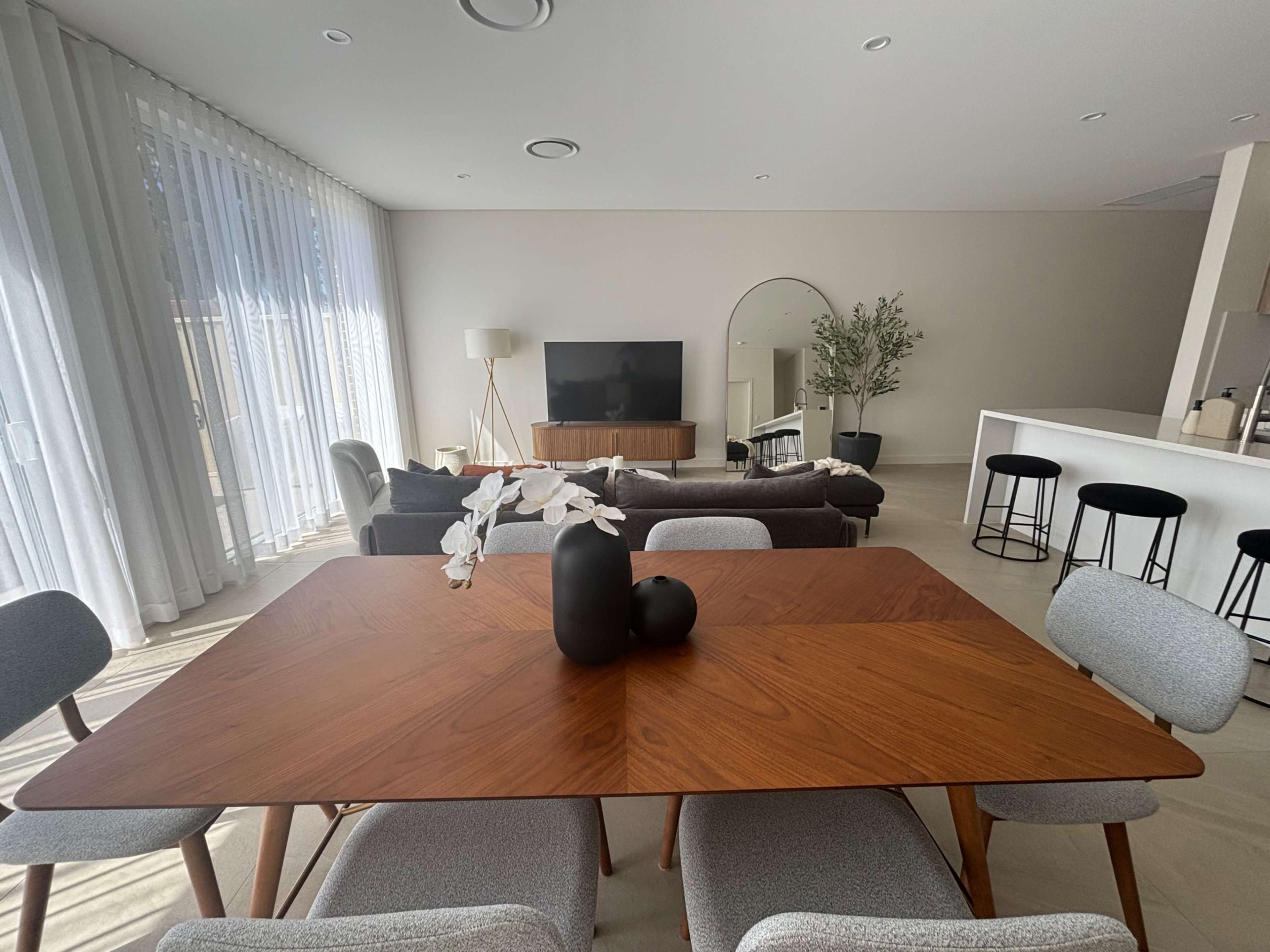 A modern living space features a wooden dining table with vases at its center, surrounded by upholstered chairs, a sofa area, and a television mounted on the wall.