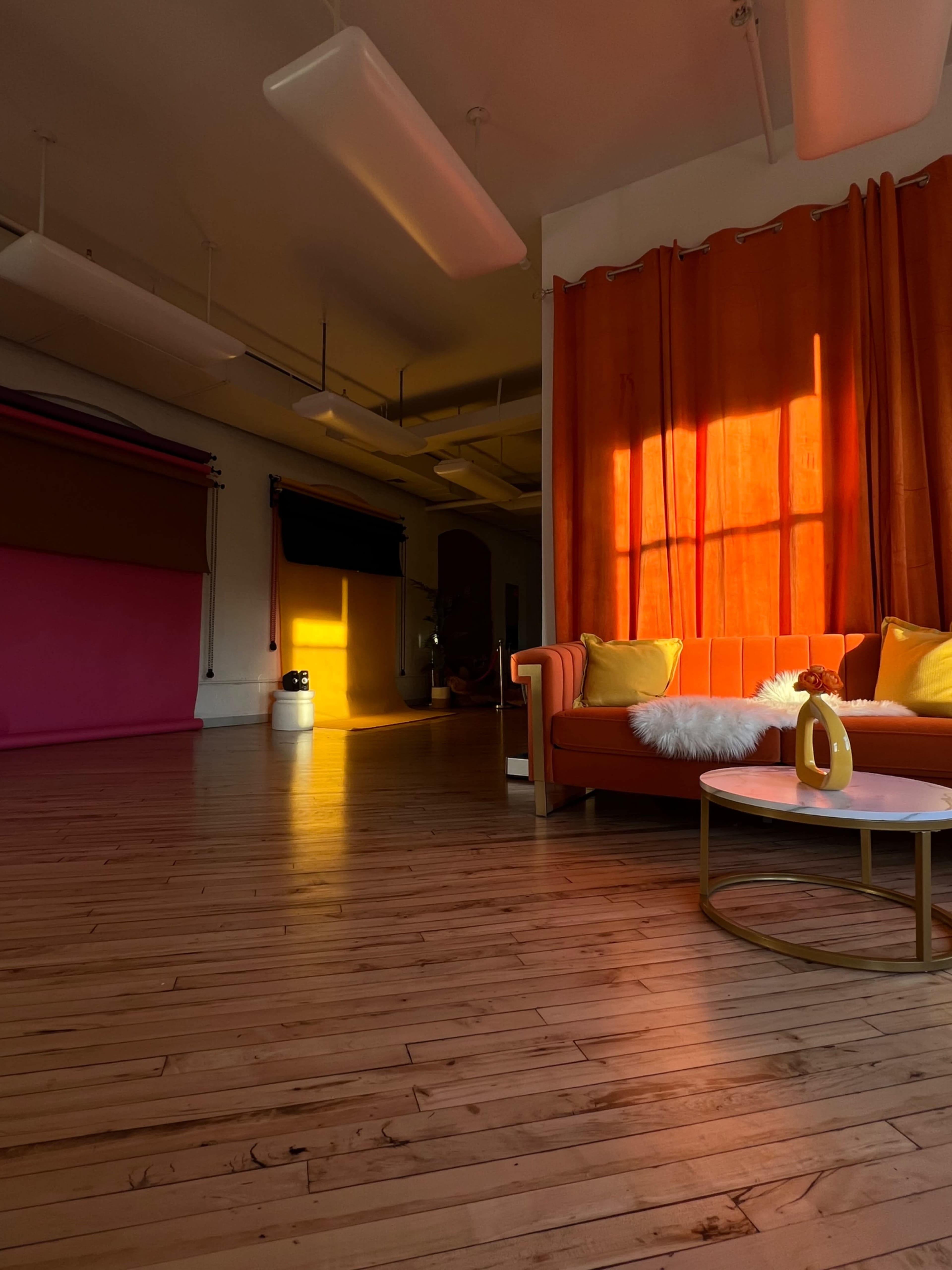 A brightly lit room features an orange curtain, a pink backdrop, a wooden floor, and a cozy orange sofa with a white pillow and a round table.
