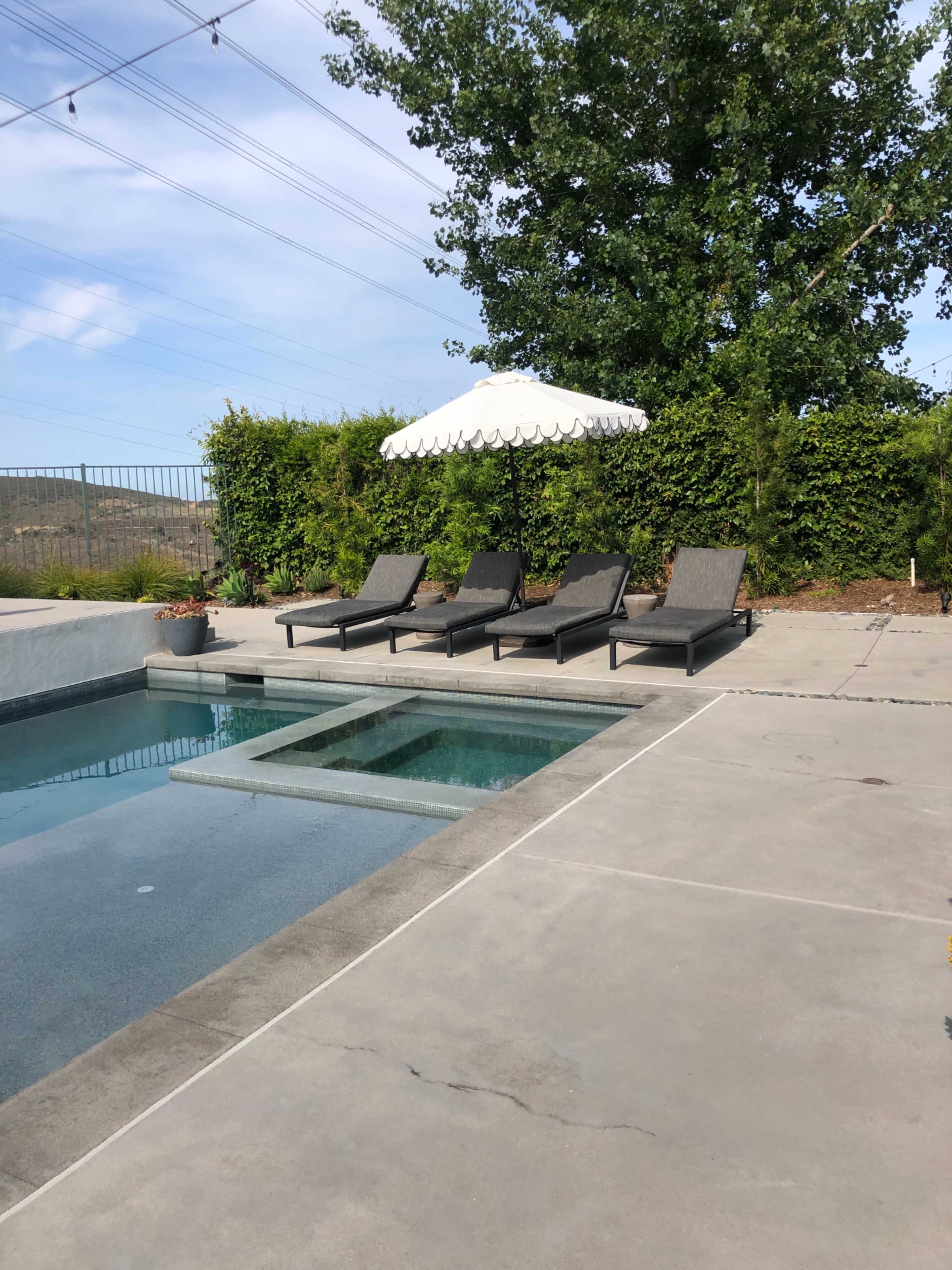 A pool area features four lounge chairs under a large umbrella beside a clear water pool.