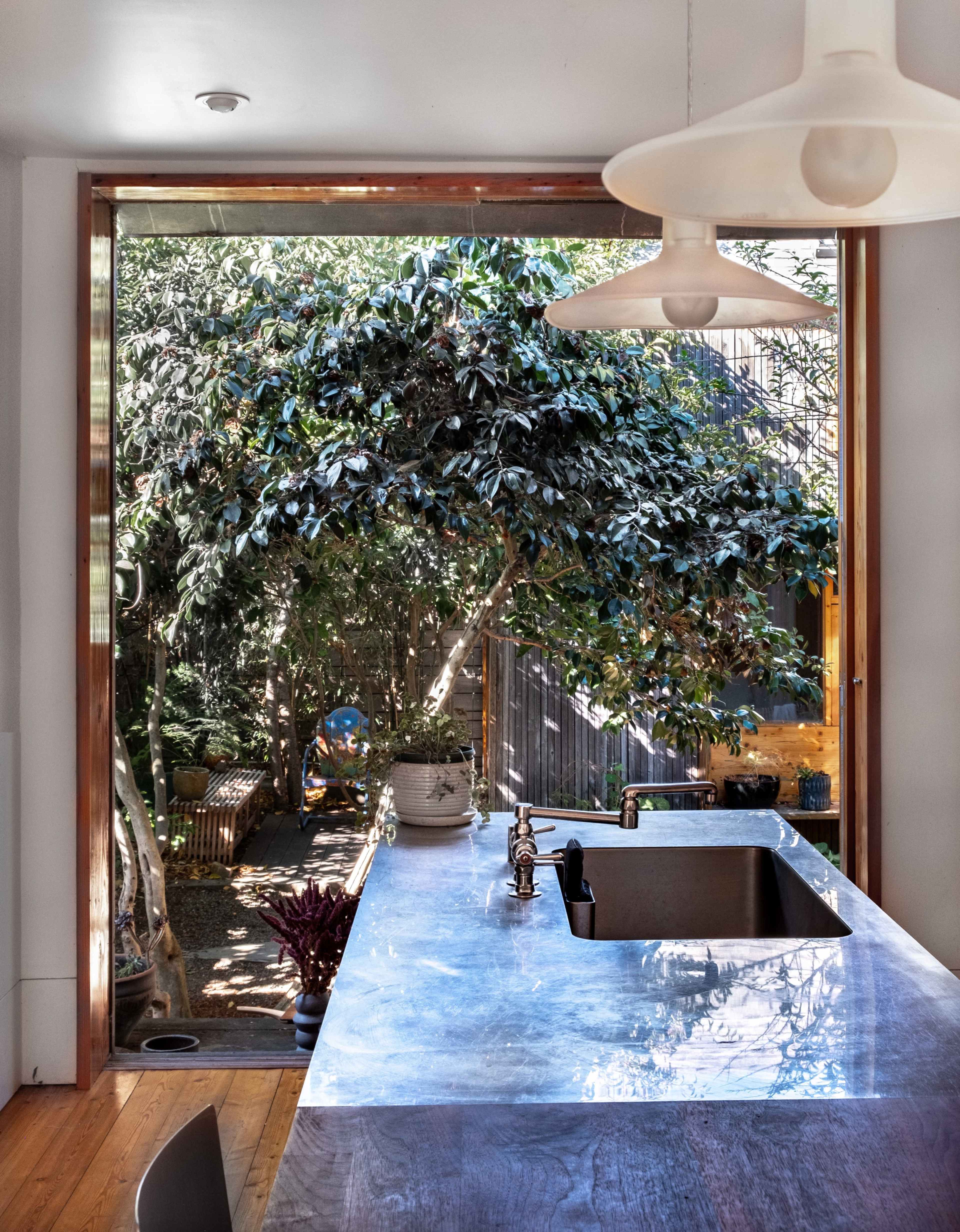 A modern kitchen with a marble countertop overlooks a garden through large windows framed in wood.