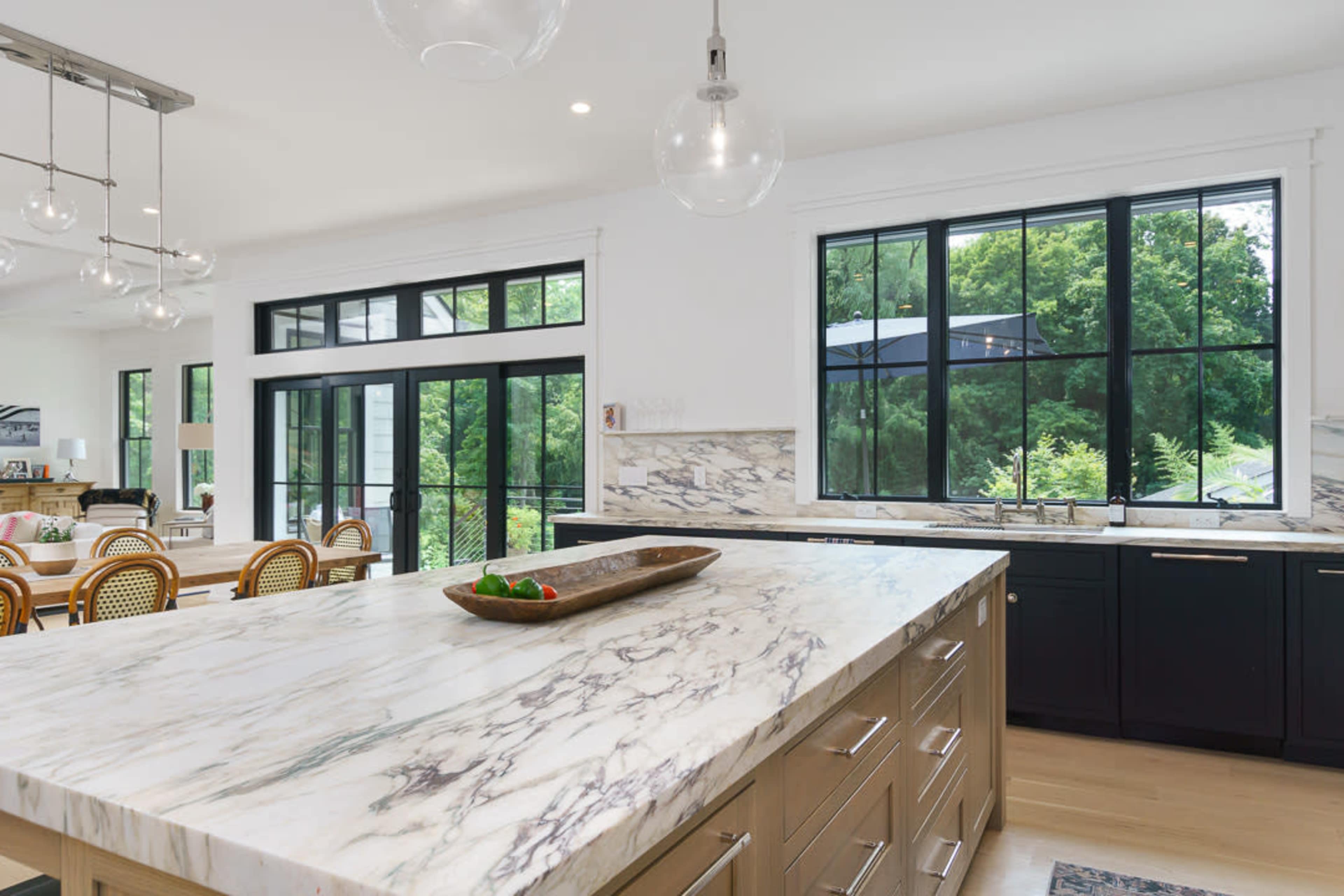 A modern kitchen features a large marble island, dark cabinetry, and large windows that offer a view of the outdoors.