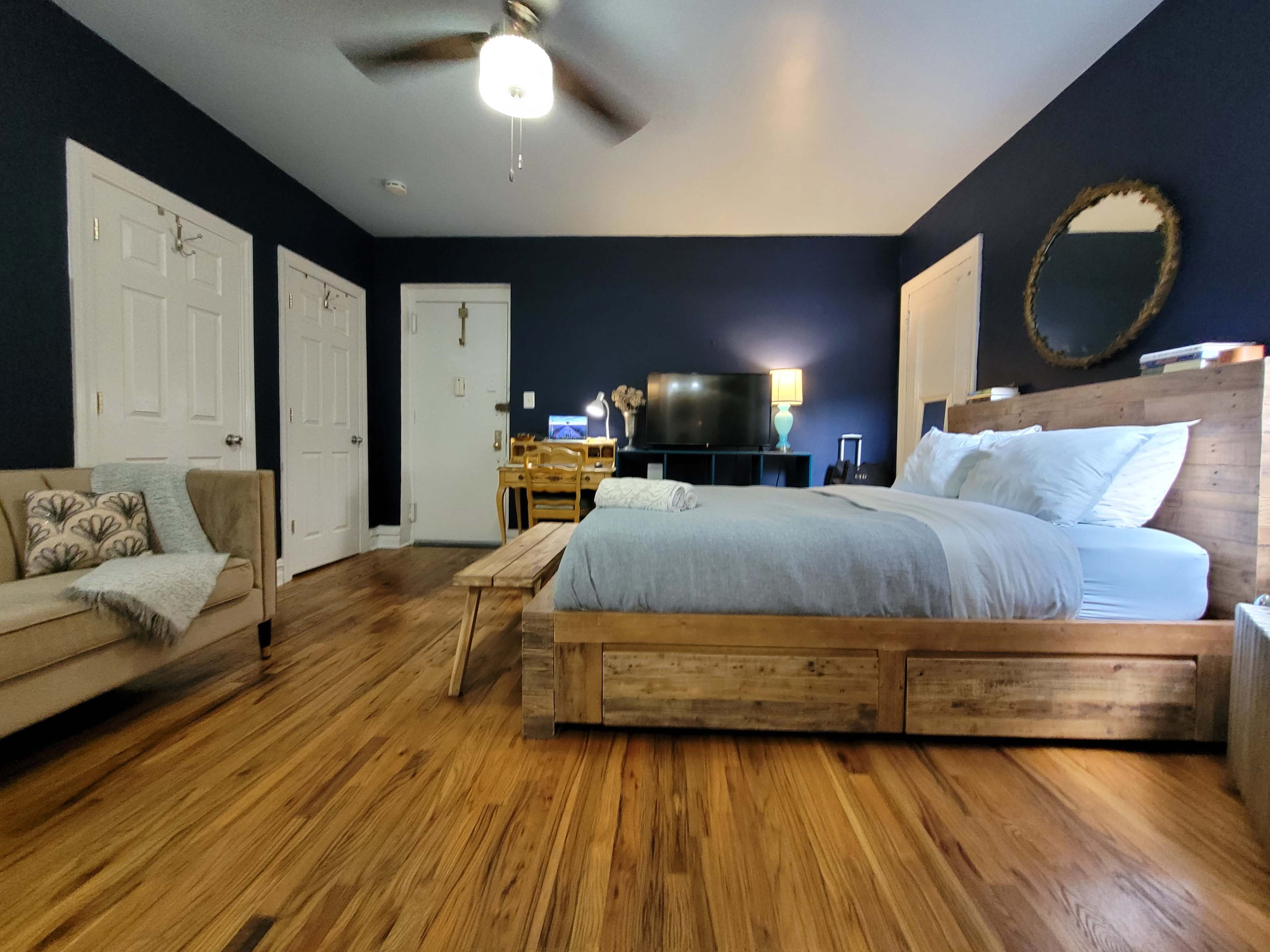 The image shows a bedroom with a wooden bed, a television on a stand, a small dining table, and a seating area, all set against dark blue walls and hardwood flooring.