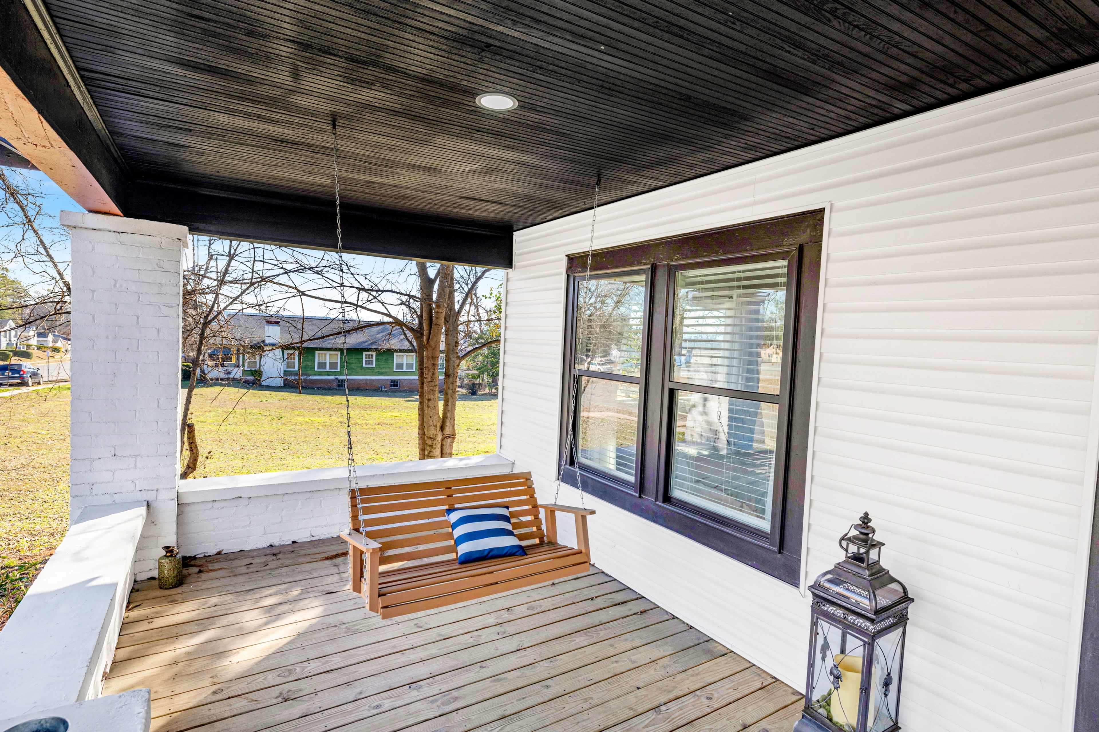 A wooden swing is suspended from the ceiling on a porch, beside a window and a lantern.
