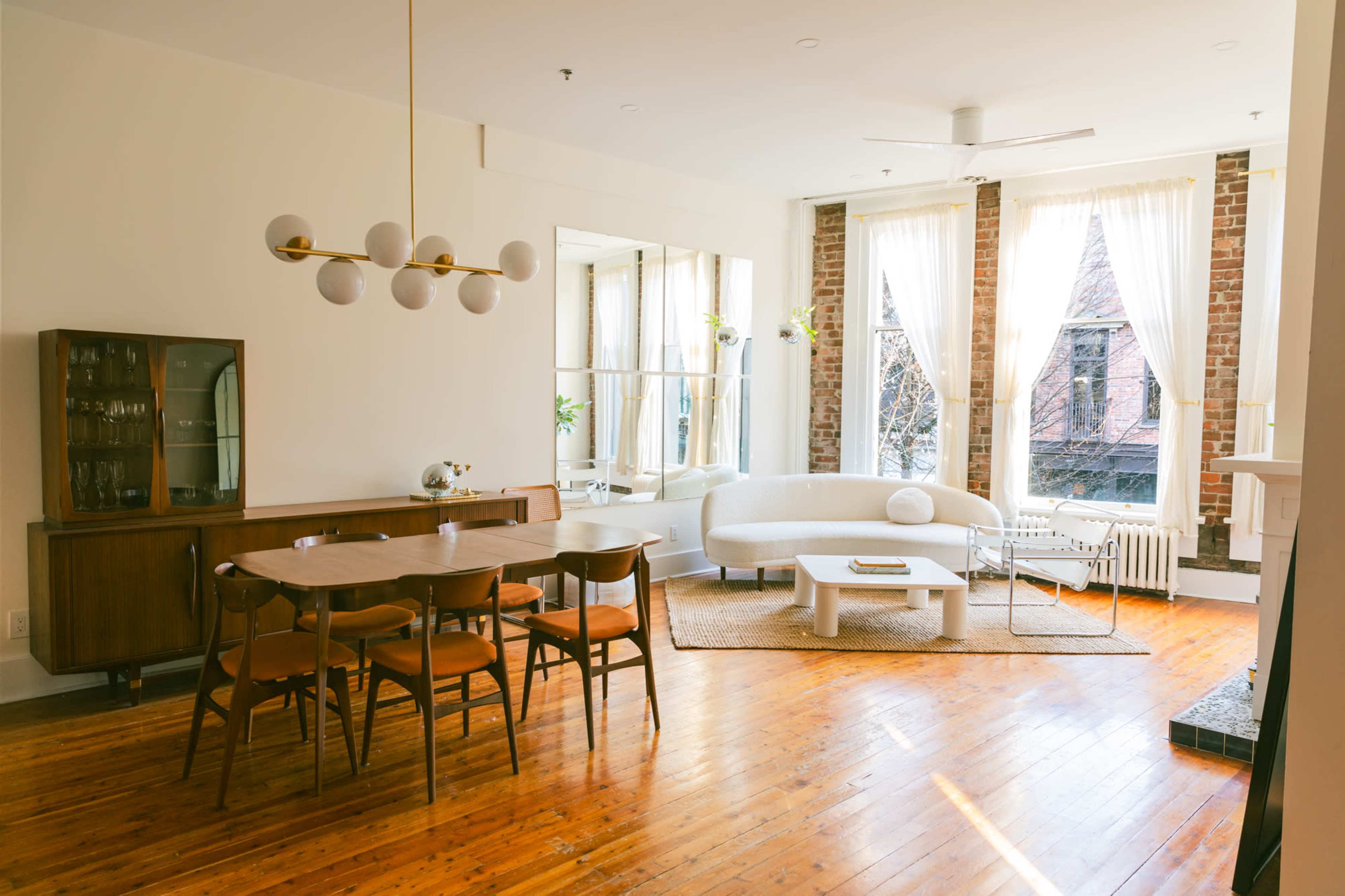 A spacious, well-lit living area featuring a dining table with wooden chairs, a modern curved sofa, and large windows that allow natural light to fill the room.