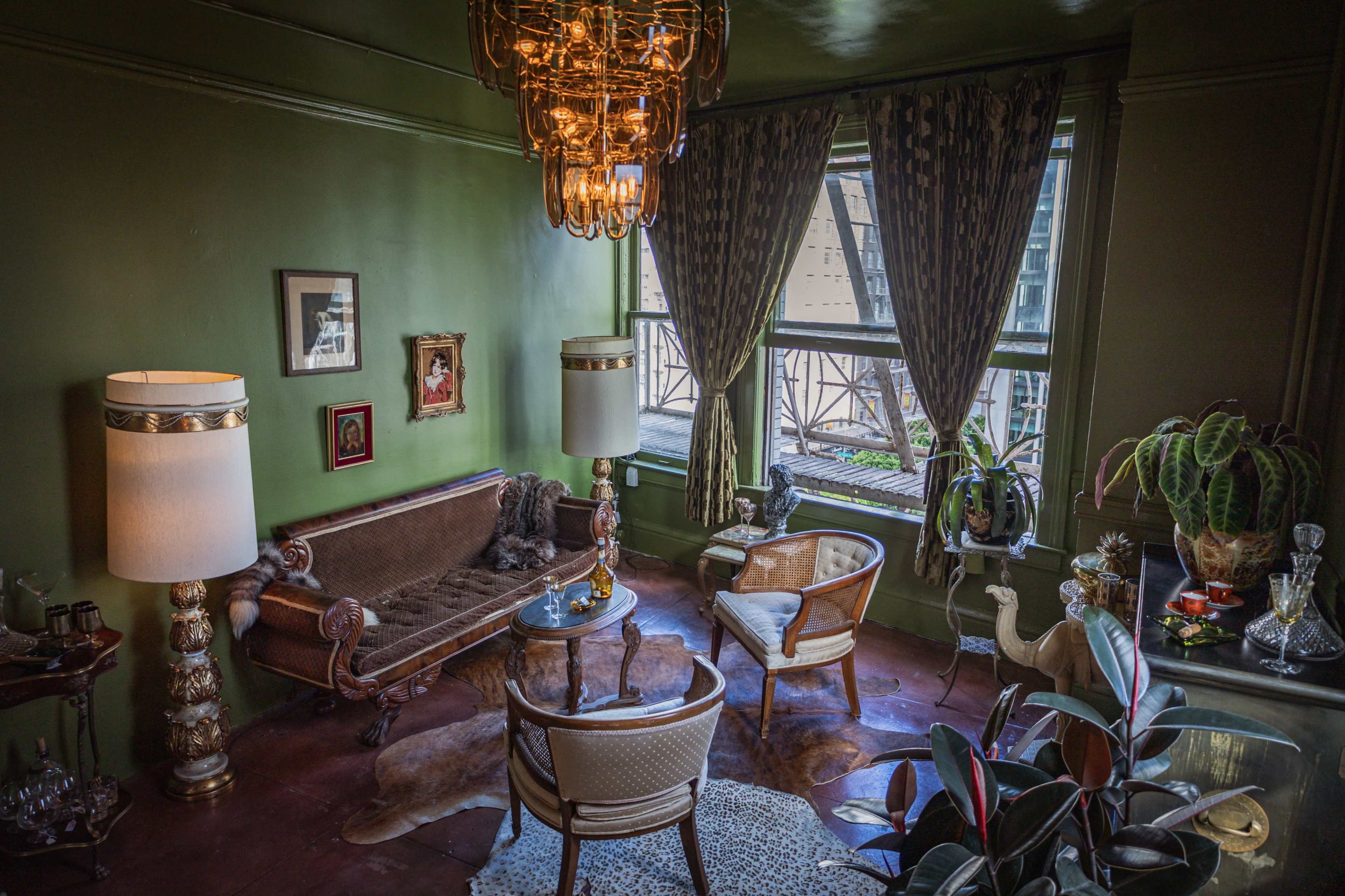 The image shows a vintage-style living room with green walls, ornate furniture, and large windows allowing natural light to enter.