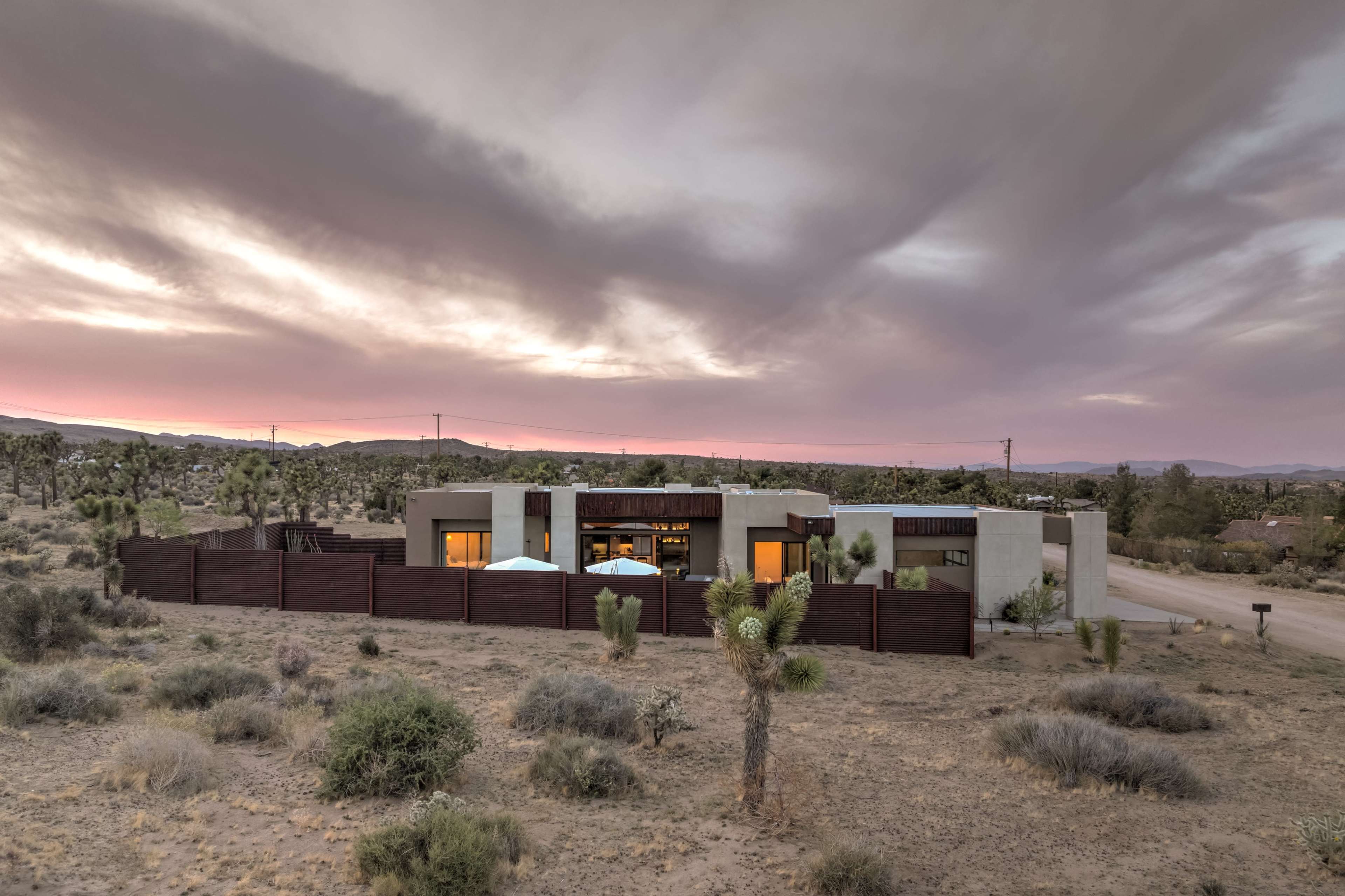 The image shows a modern house surrounded by desert landscape under a cloudy sky at dusk.