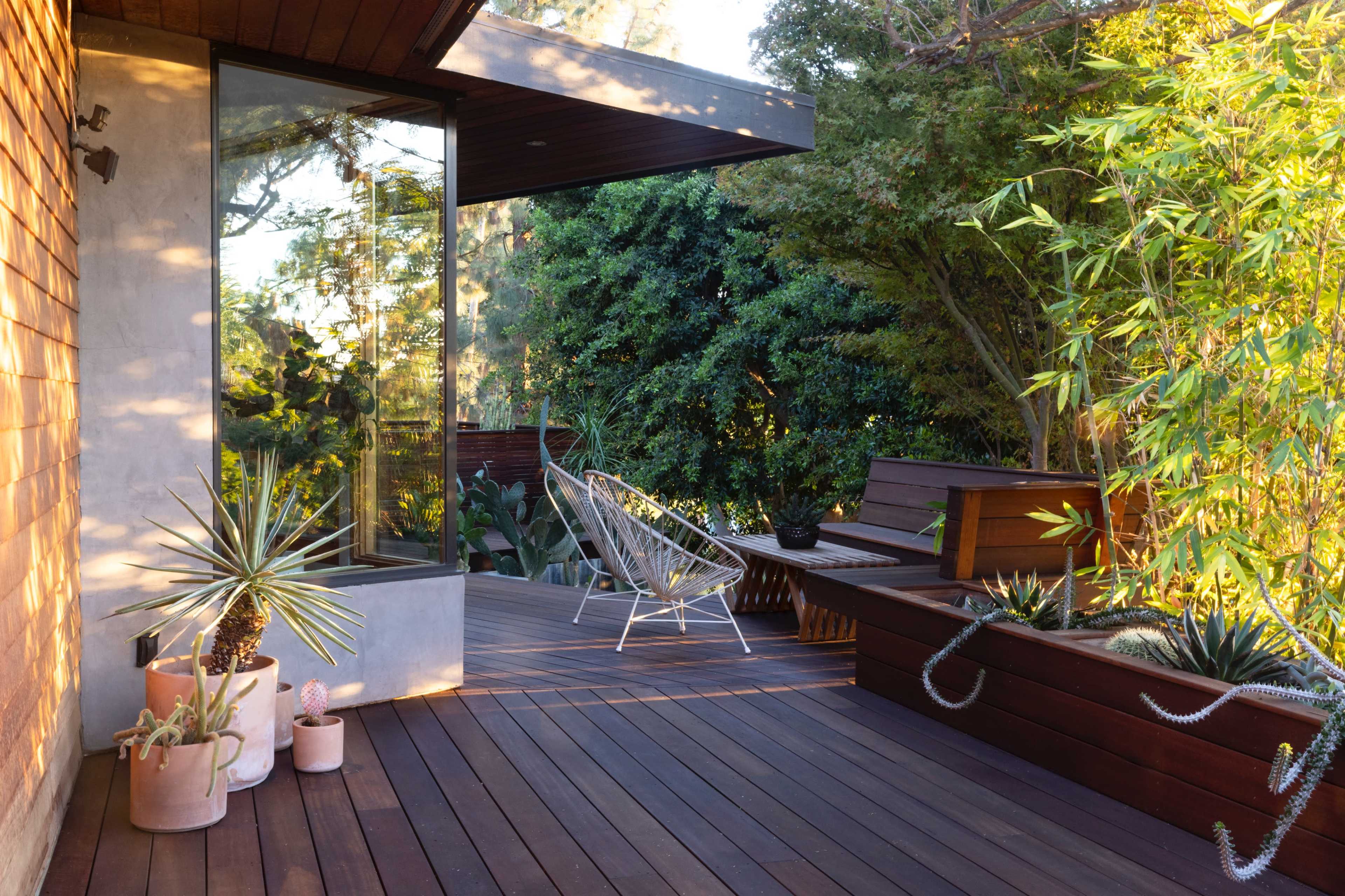 The image shows a wooden deck area adjoining a glass-walled structure, featuring potted plants, outdoor seating, and surrounding greenery.