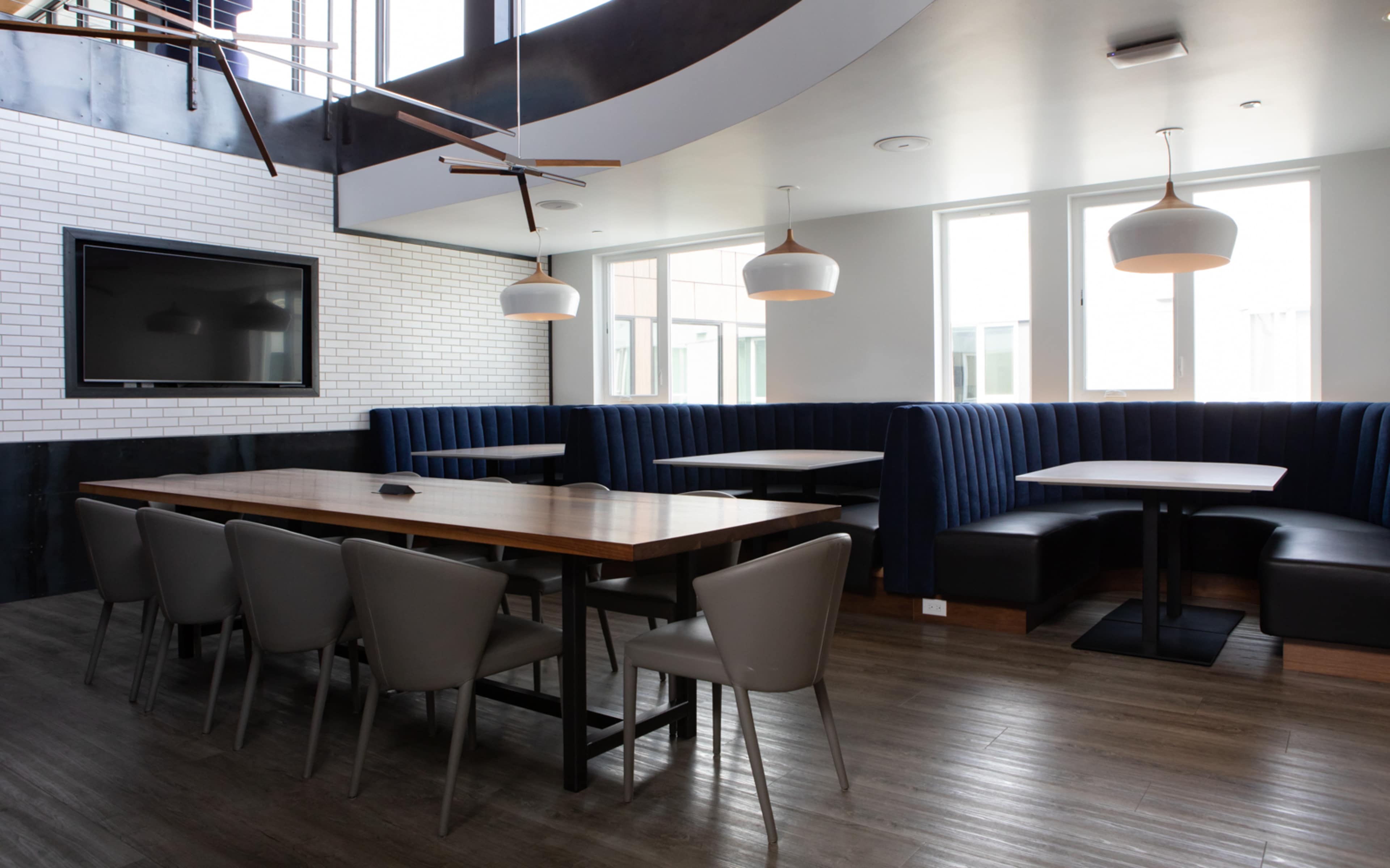 A modern dining area with a long wooden table, gray chairs, and blue upholstered booths, along with large windows and a flat-screen TV on the wall.