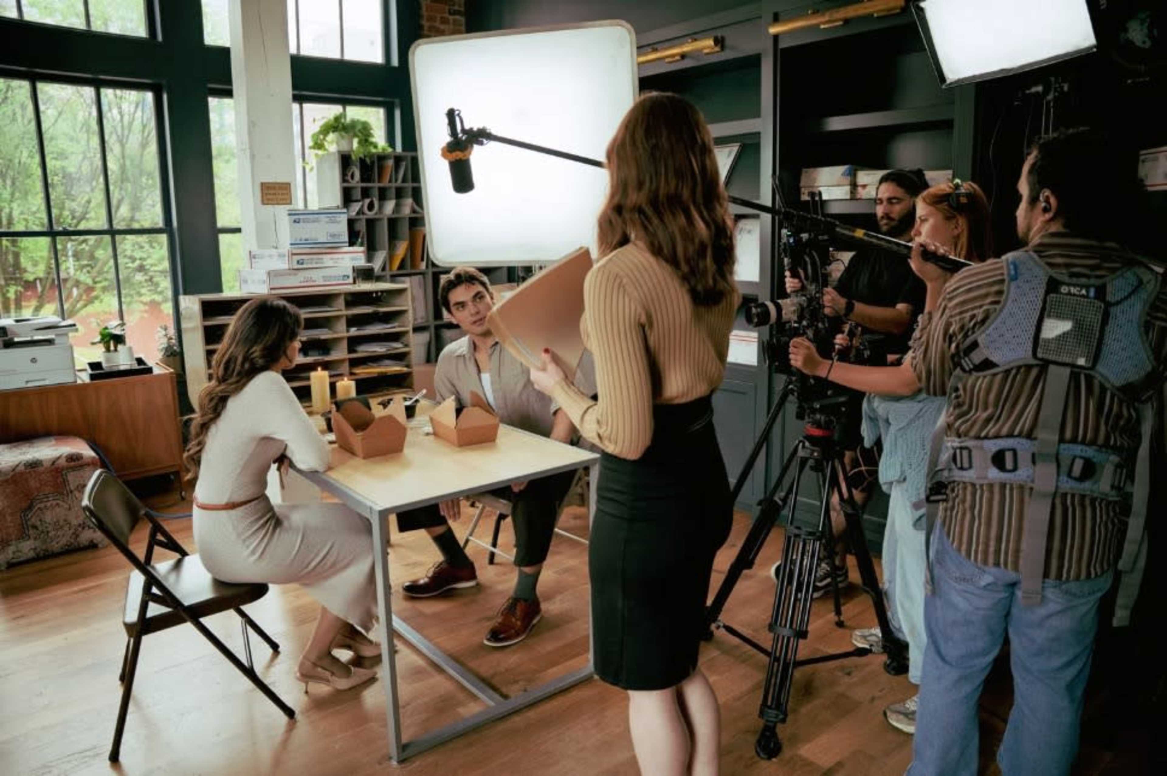 A film crew sets up a scene in a studio featuring two actors seated at a table with takeout containers.