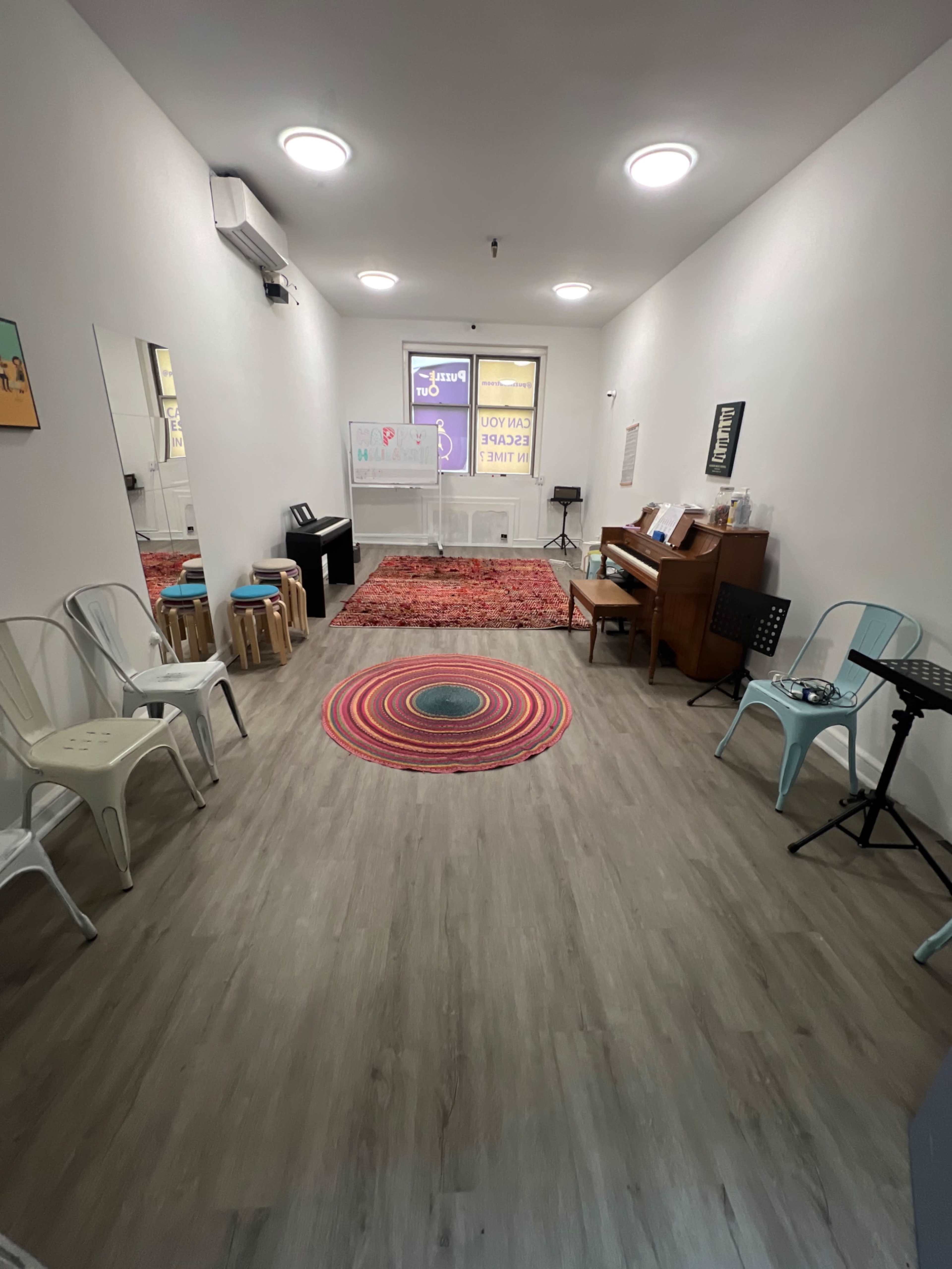 The image shows a bright, empty room with wooden flooring, a piano against one wall, chairs arranged around a circular rug, and colorful wall art.