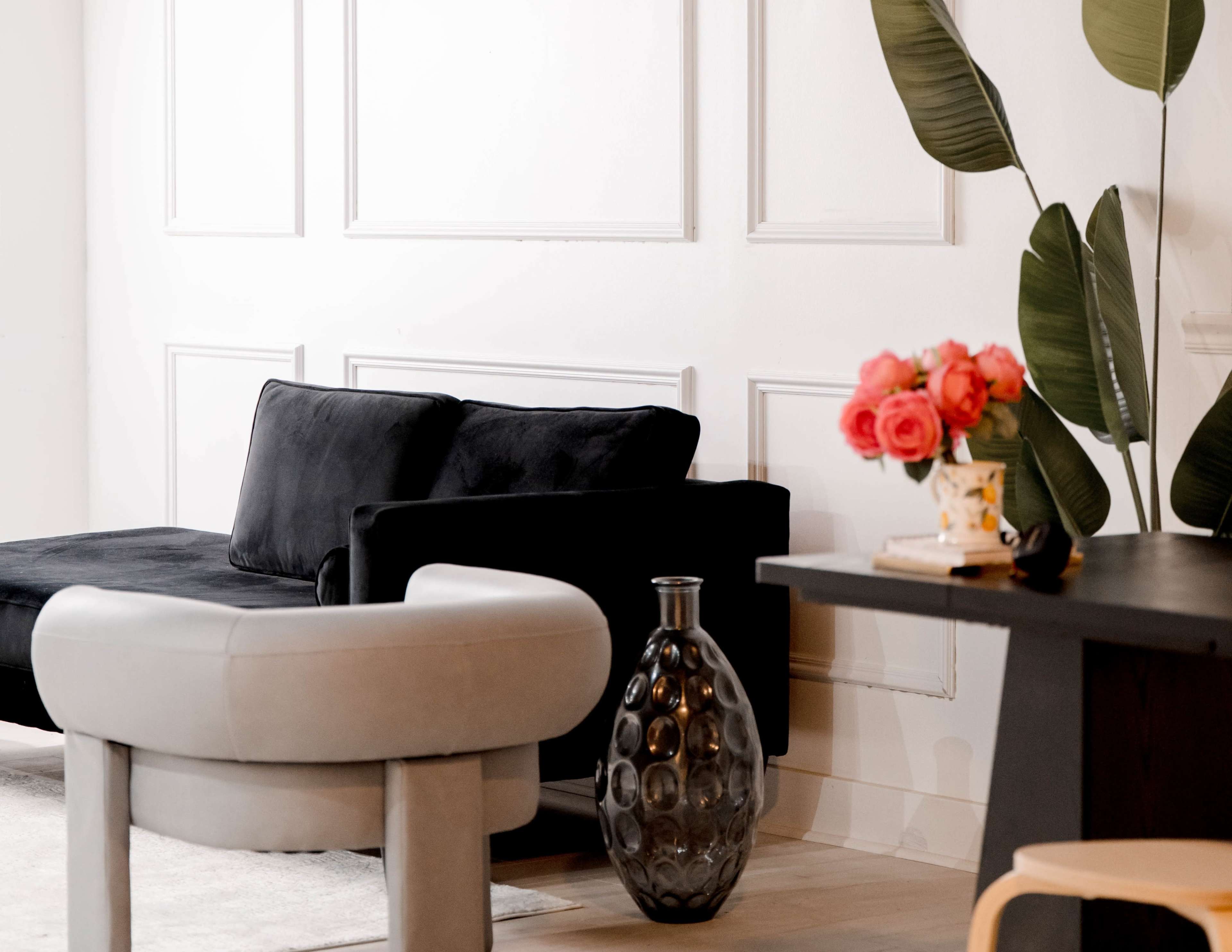 The image shows a modern interior featuring a black sofa, a curved gray chair, a decorative black vase, and a table with a flower arrangement.