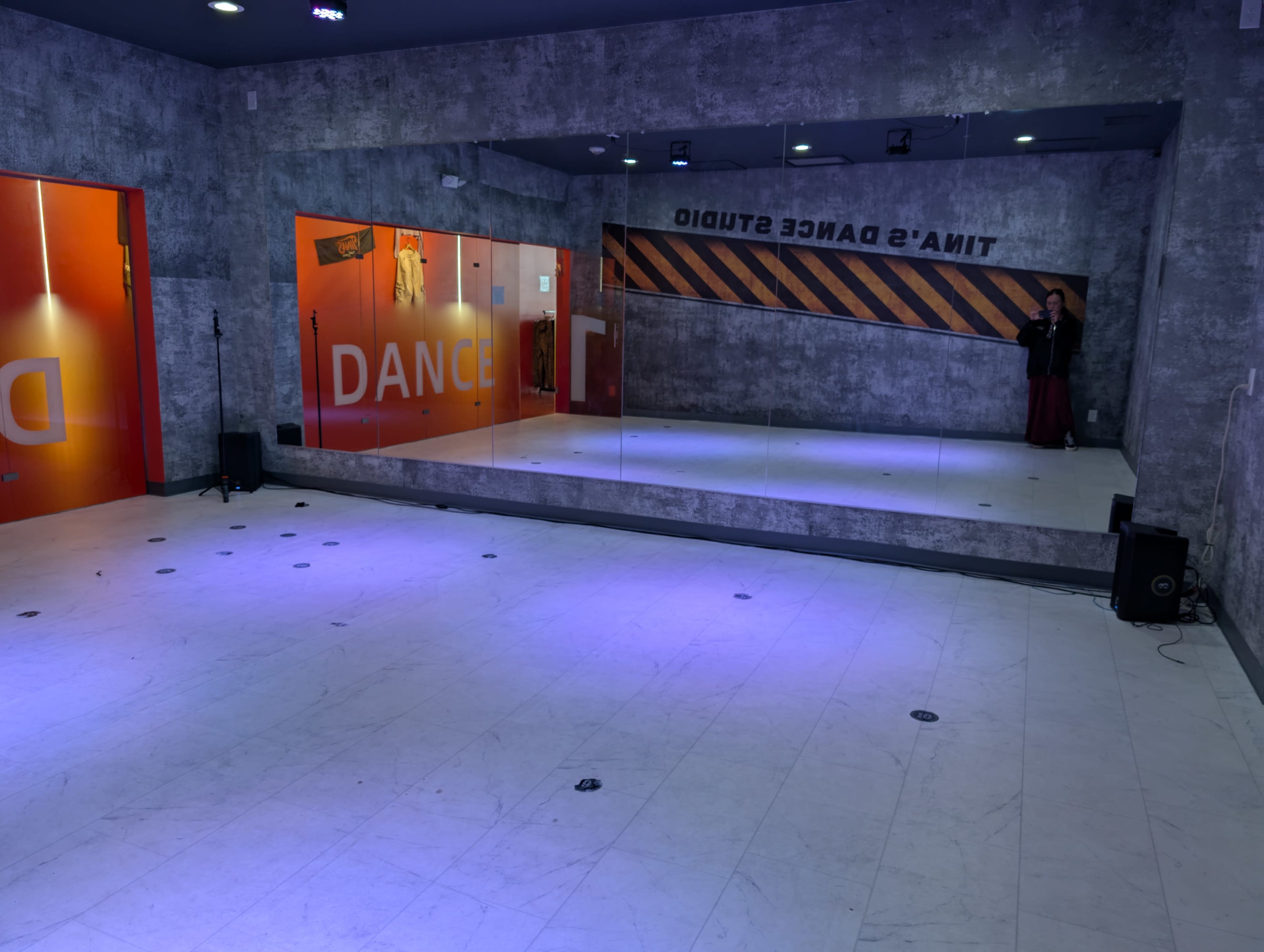 The image shows a dance studio with mirrored walls, gray textured walls, and orange accents.