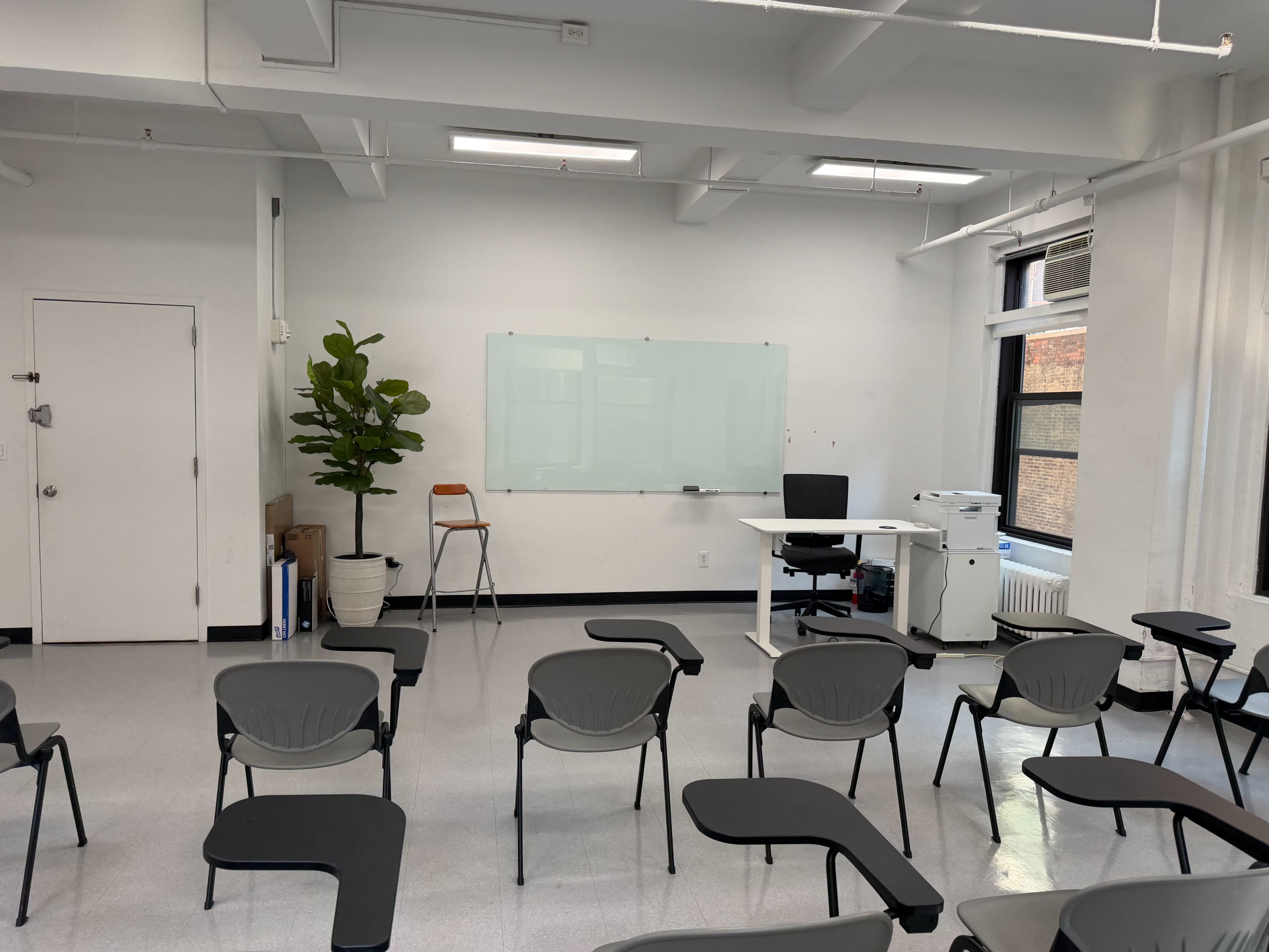 The image shows a spacious, empty classroom with rows of chairs facing a whiteboard, a desk, and a printer.
