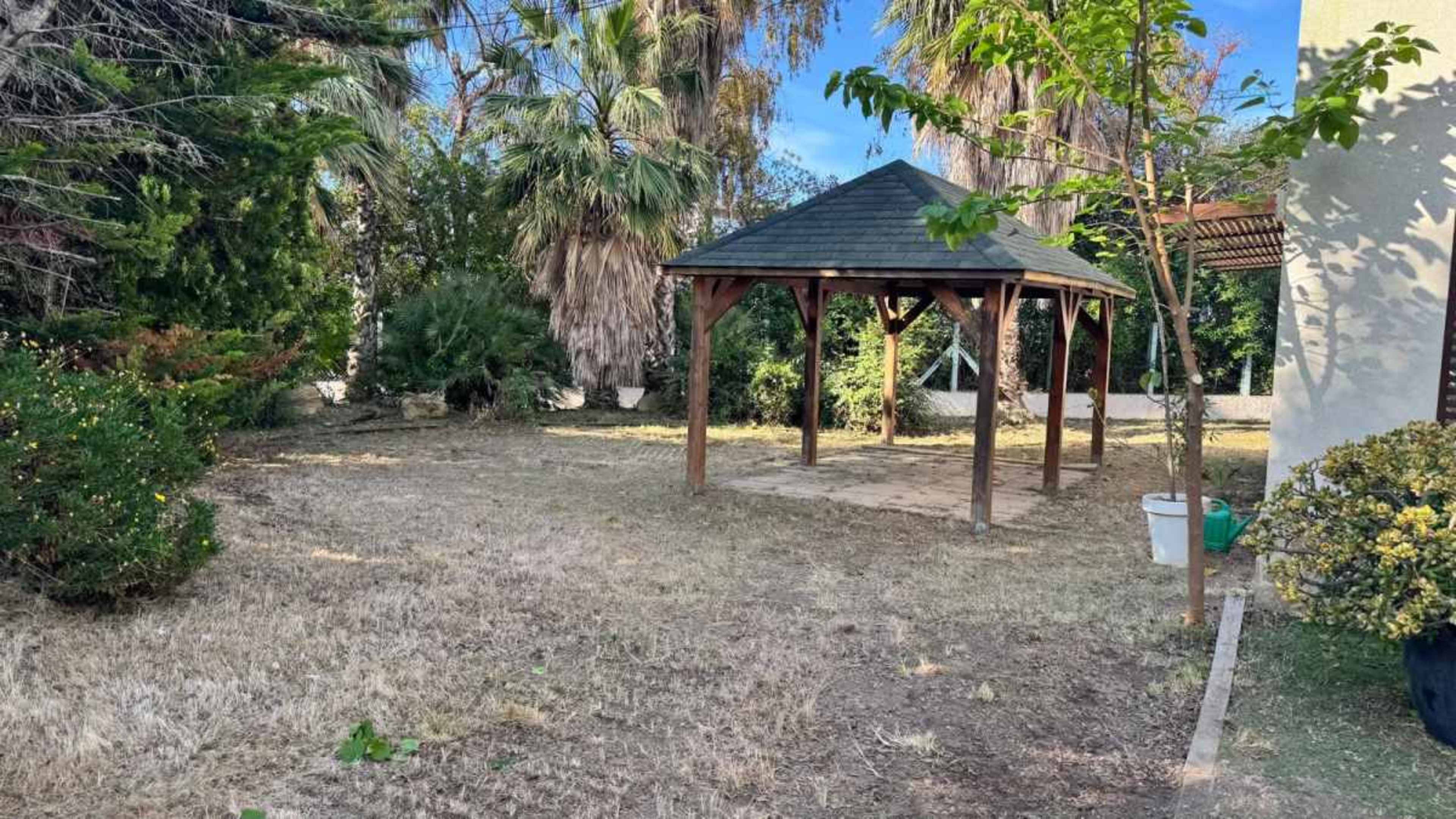 The image shows a wooden gazebo in a grassy area surrounded by trees and shrubs.