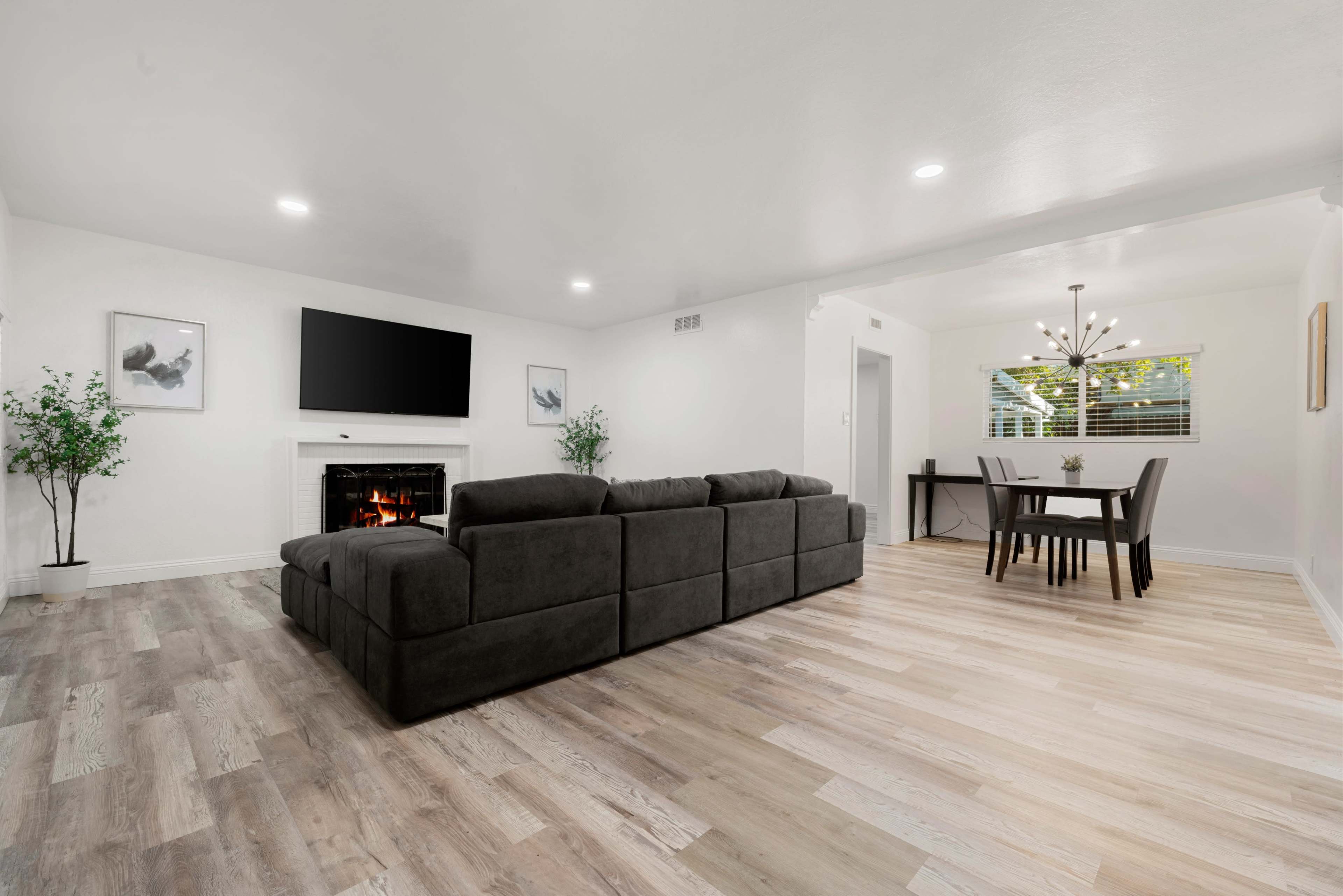 A modern living room with a dark sectional sofa, a fireplace, a large wall-mounted television, and a dining area with a table and chairs.