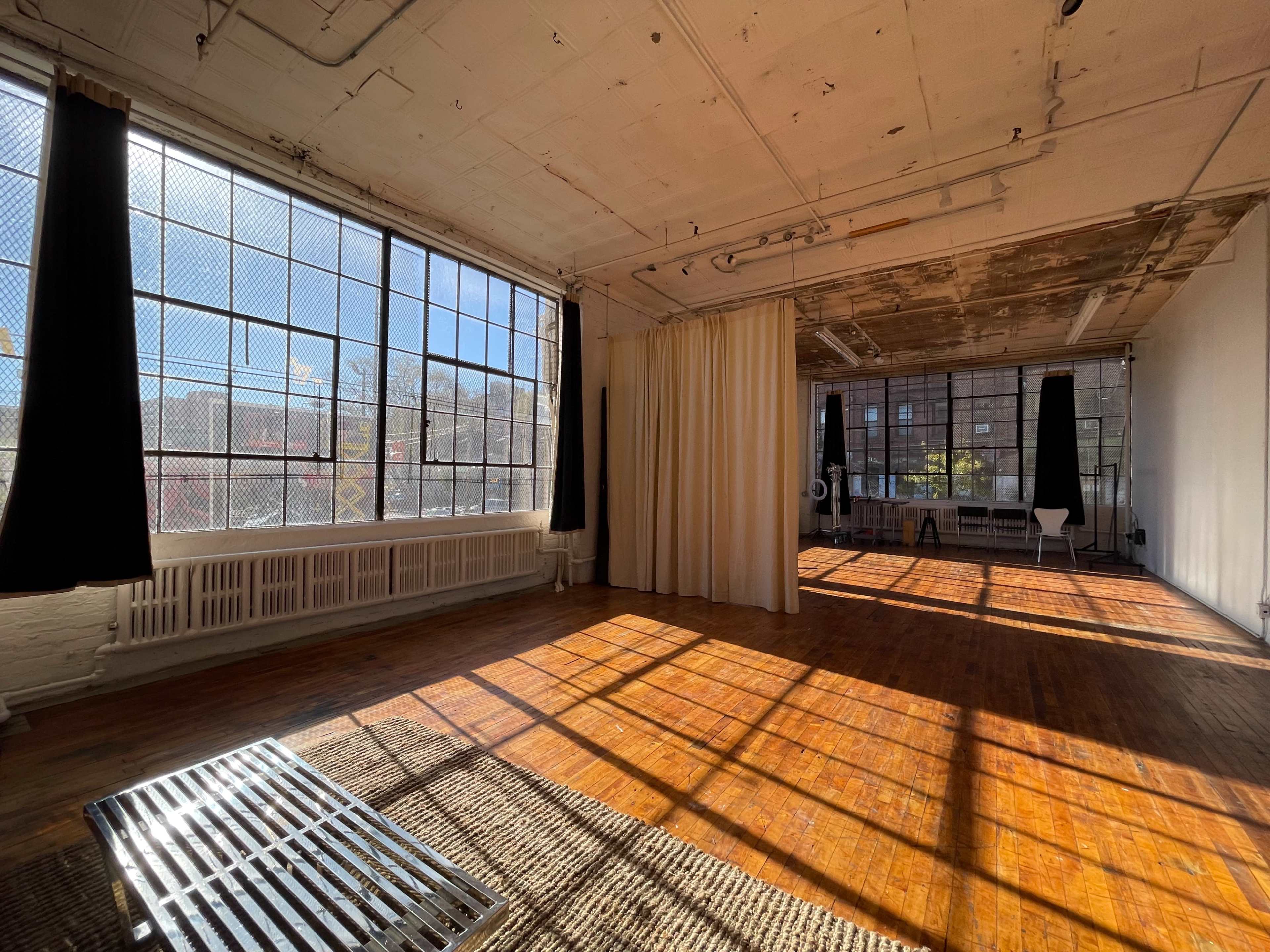 A sunlit, spacious room with large windows, wooden floors, and light-colored walls, featuring a seating area with a bench and curtains.