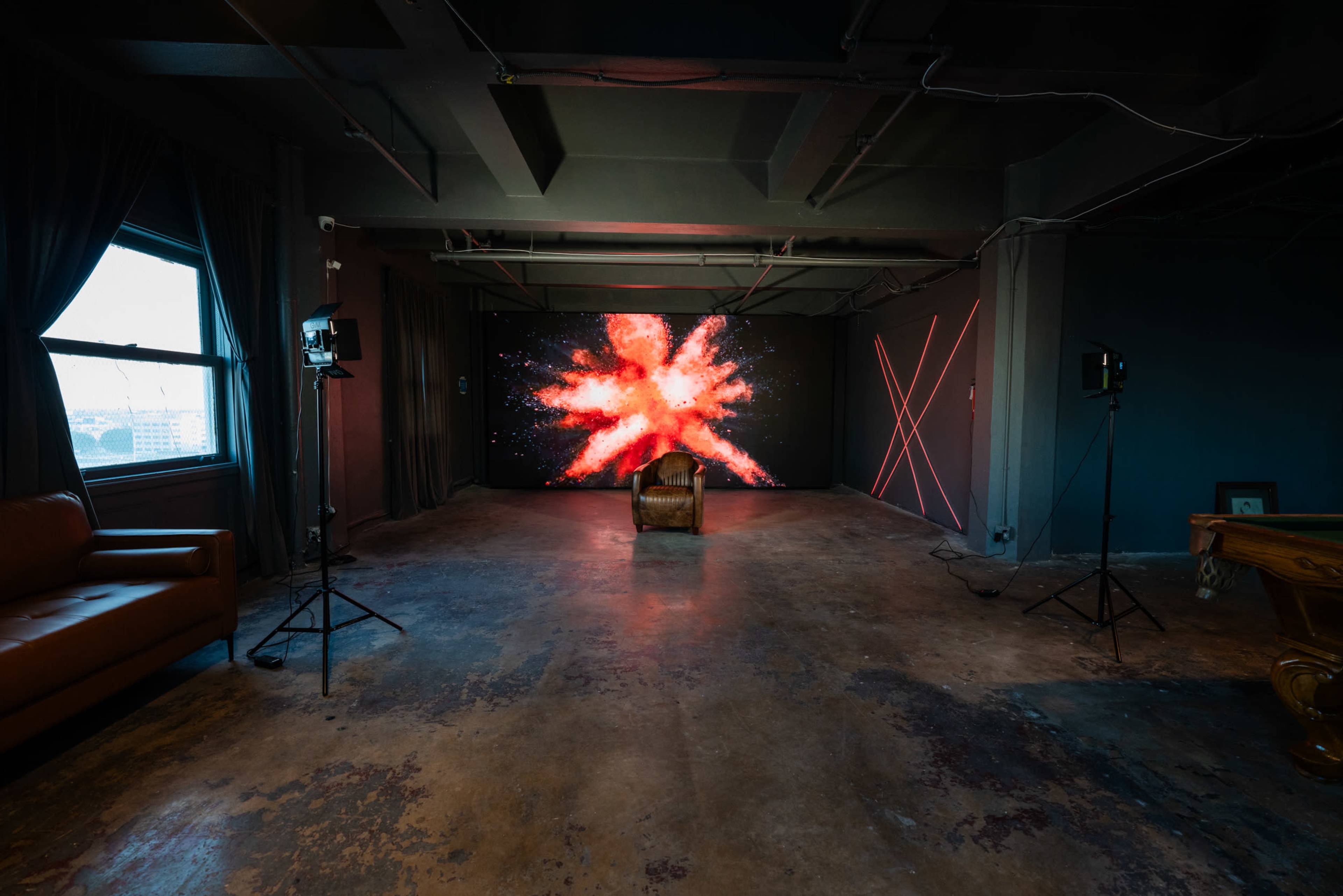 A large, brightly lit abstract display is set against a dark wall in an industrial-style room with a single chair positioned in the center.