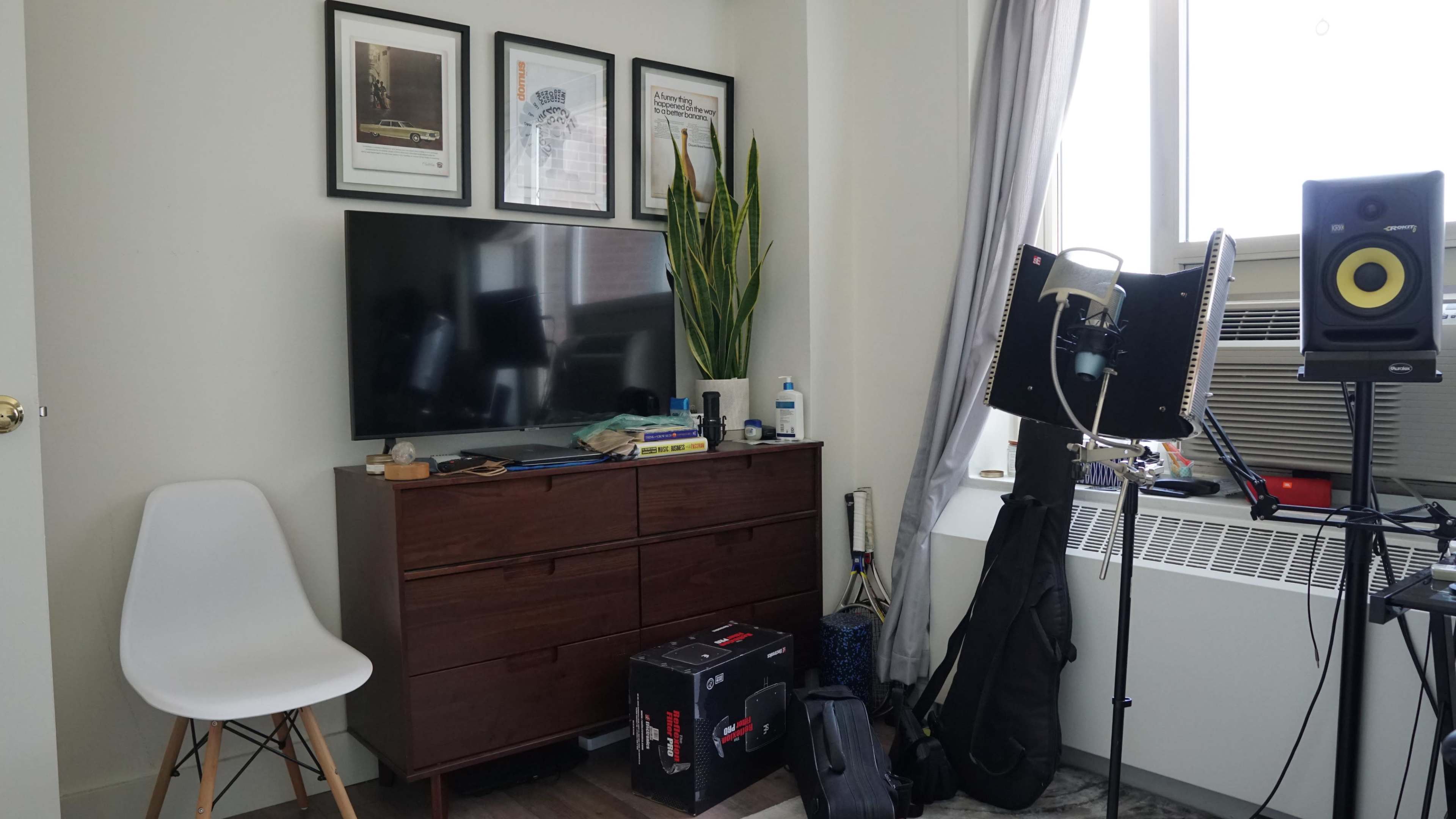 The image shows a corner of a room featuring a television on a wooden dresser, a plant, an air conditioning unit, and musical equipment.
