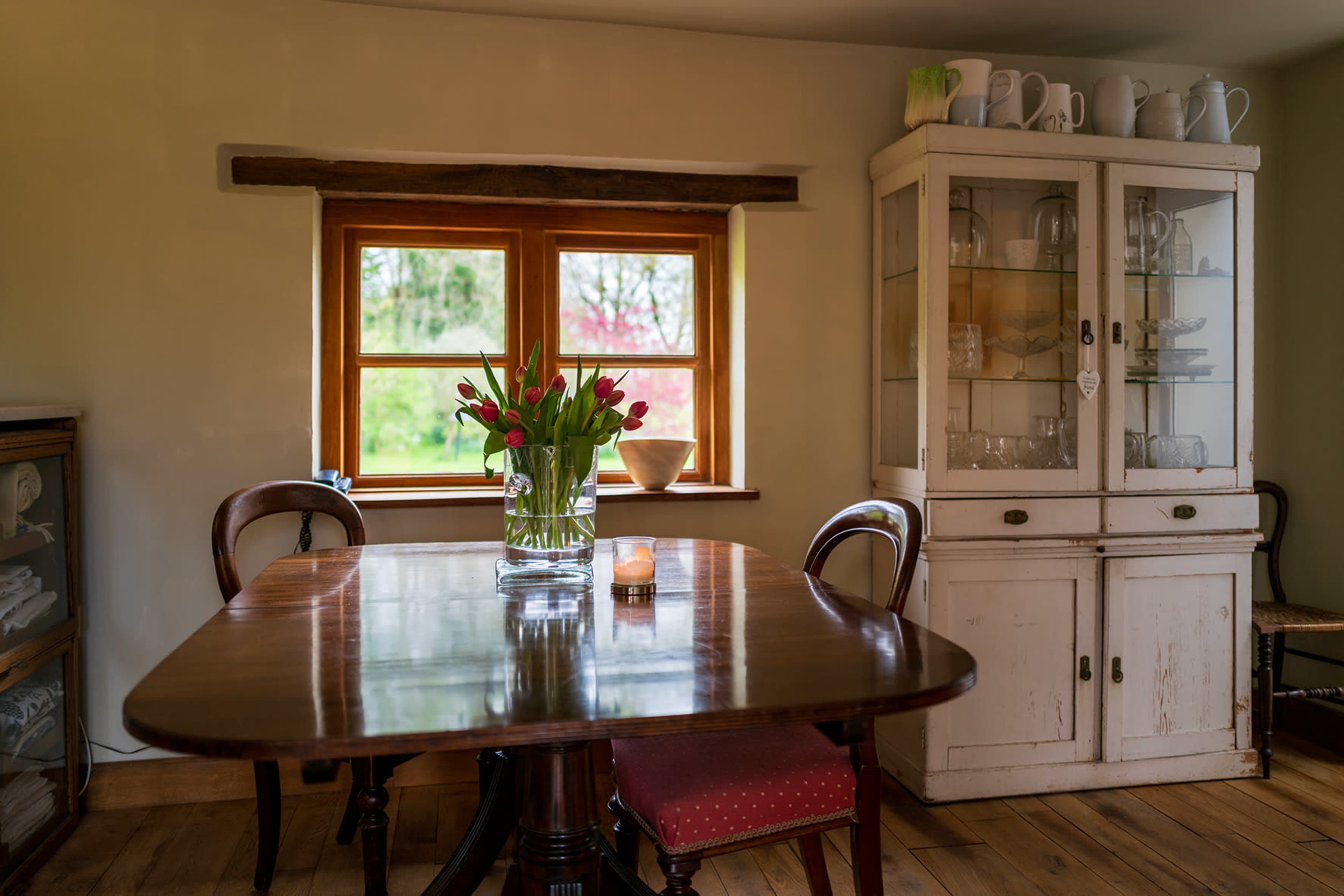 A table set with a vase of flowers next to a window and a glass-fronted cabinet displaying dishes.