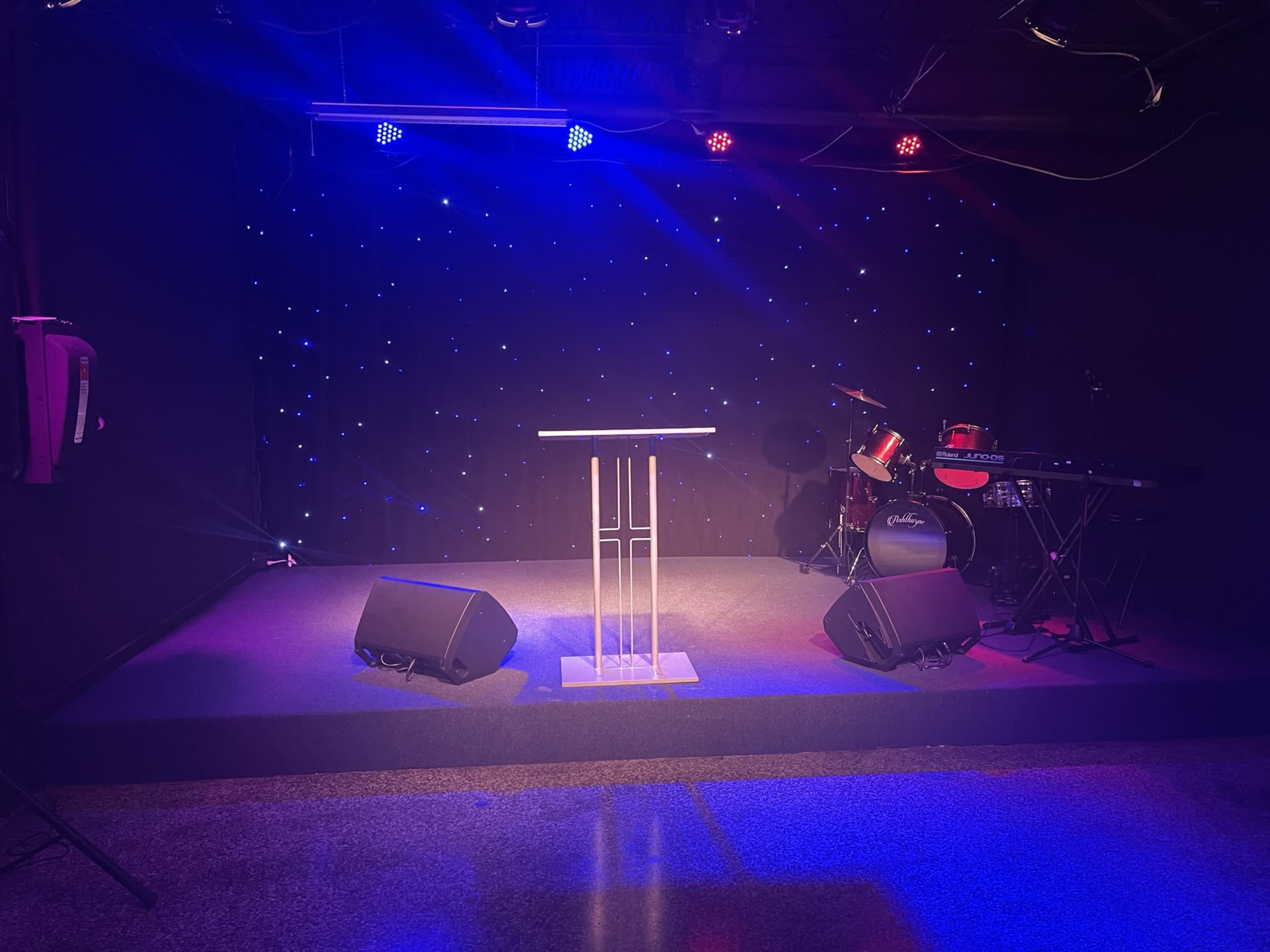 A performance stage featuring a microphone stand, a drum set, a keyboard, and illuminated by colored lights against a black backdrop with twinkling stars.