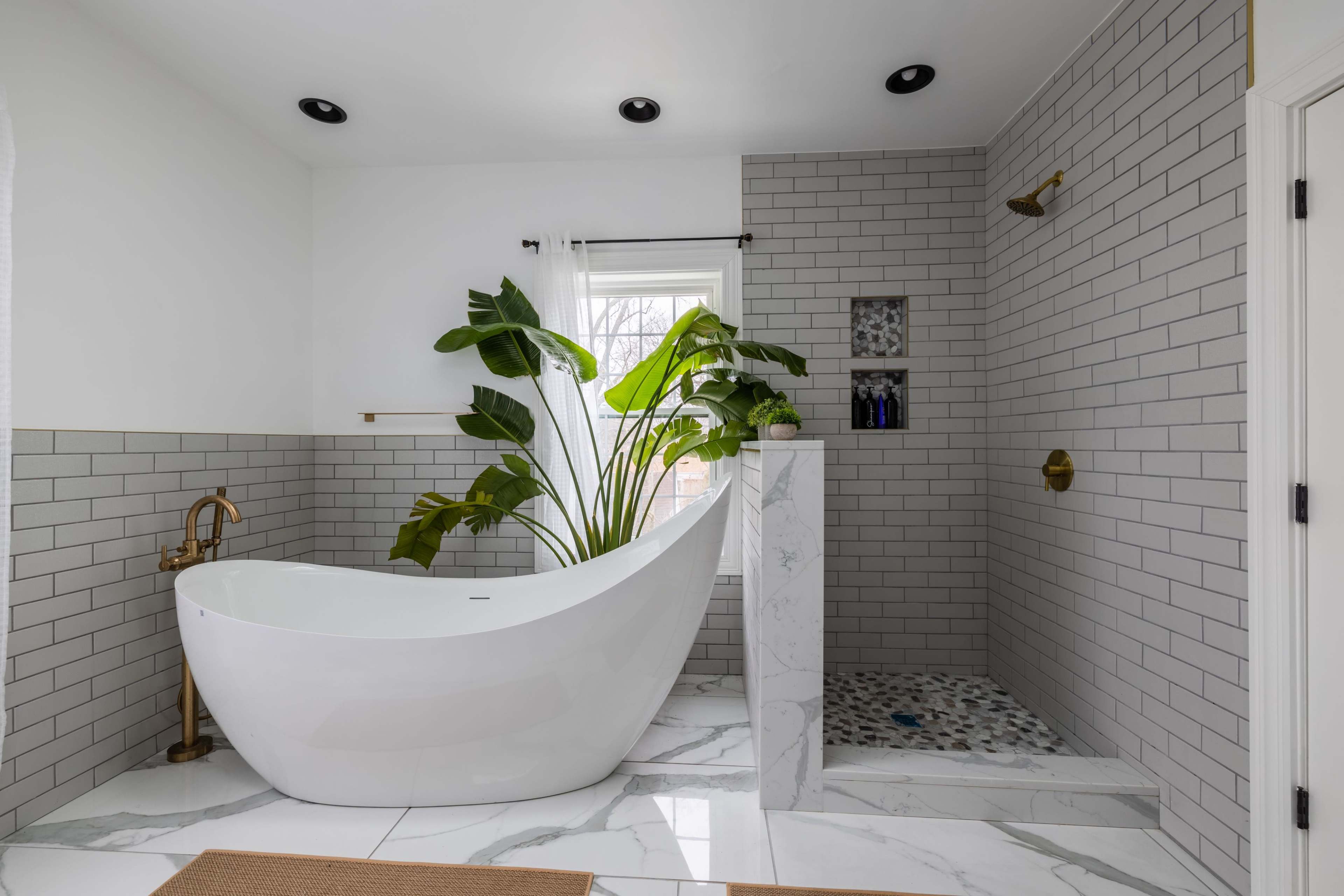 A modern bathroom featuring a freestanding bathtub, a shower area with a pebble floor, and a large plant near a window.