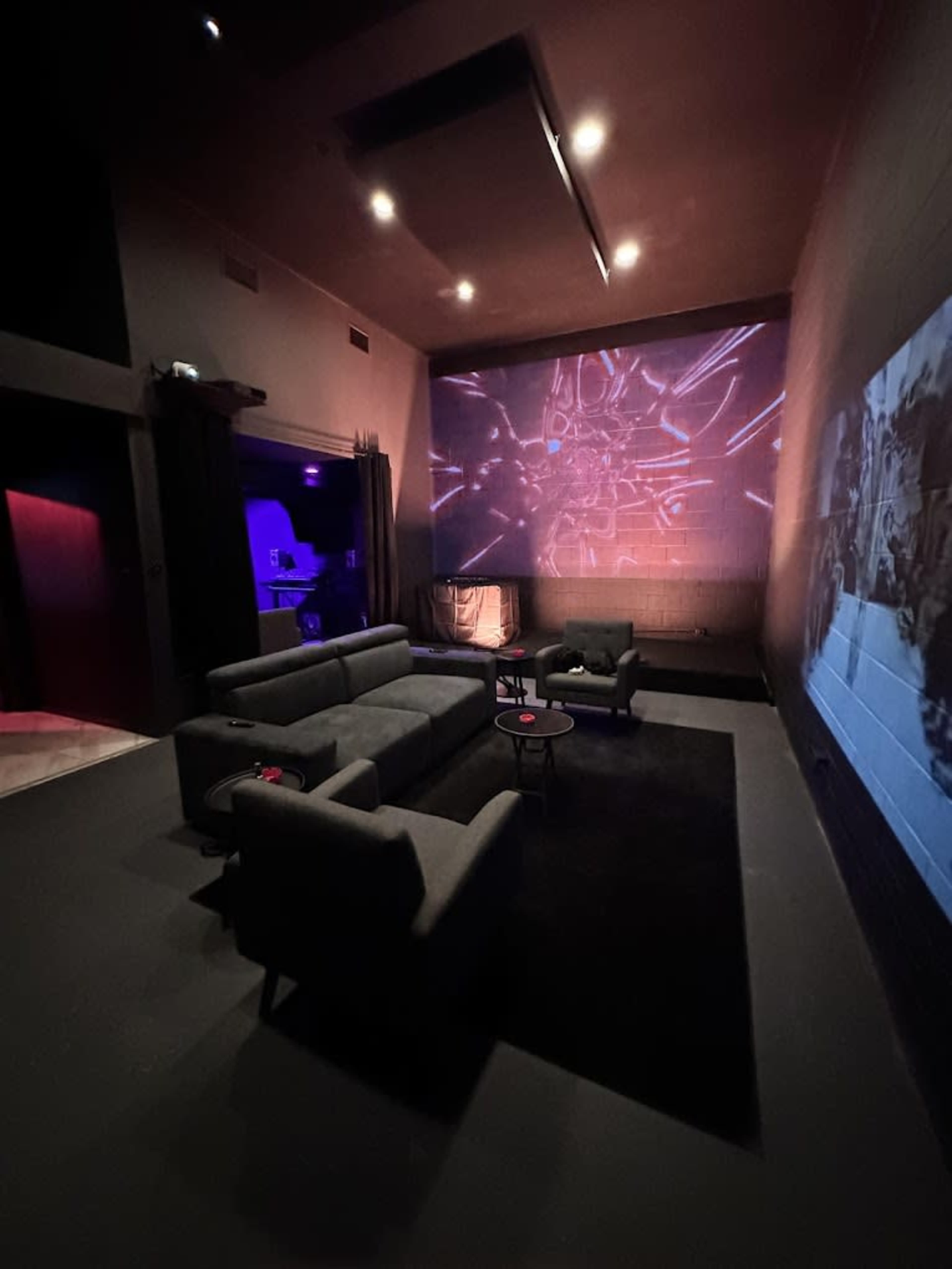 The image shows a dimly lit lounge with gray sofas, a low table, and a wall displaying a bright projection.