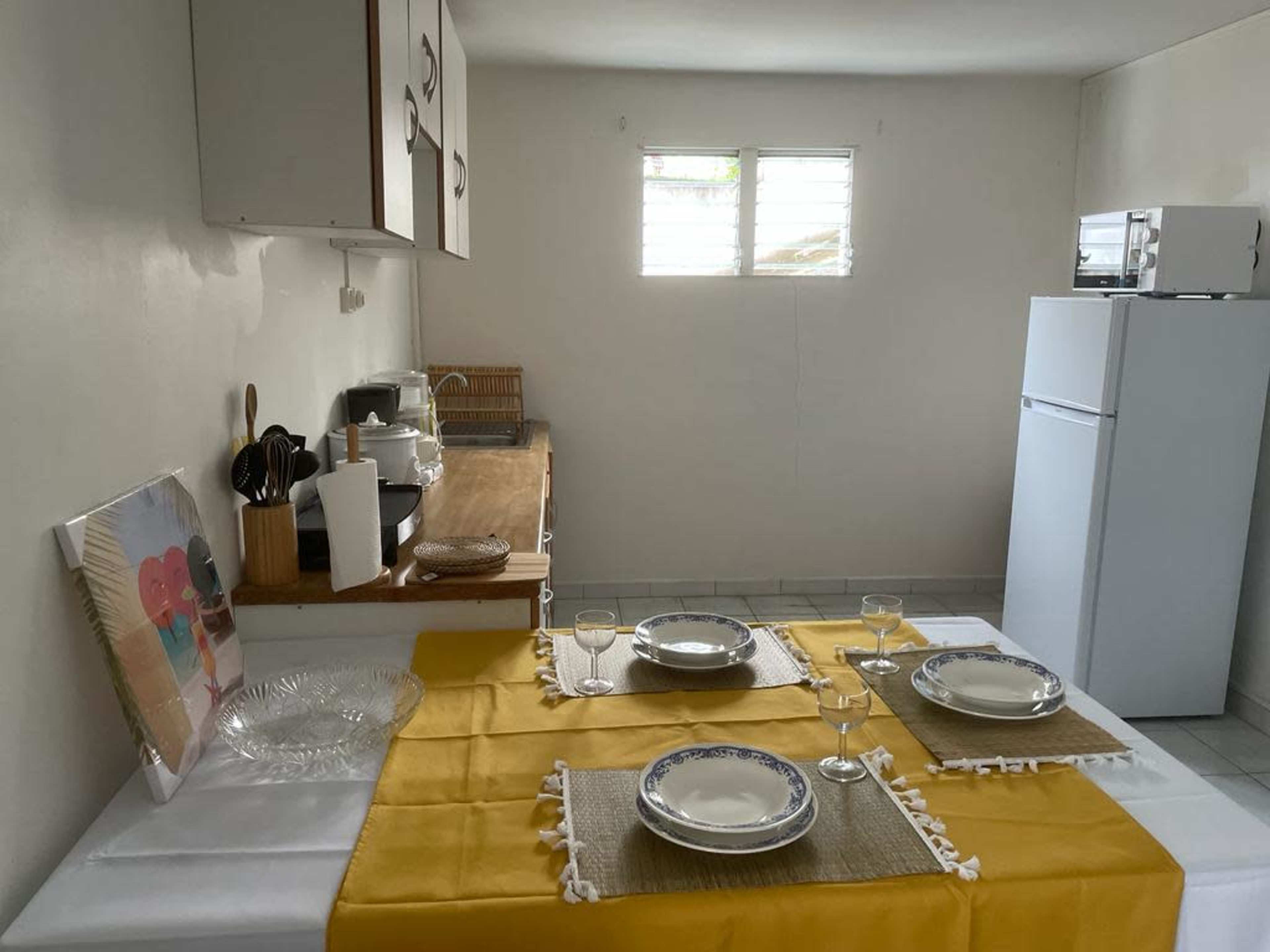 A simple kitchen setup with a wooden table set for four, featuring yellow tablecloths and plates, along with a refrigerator and a window in the background.