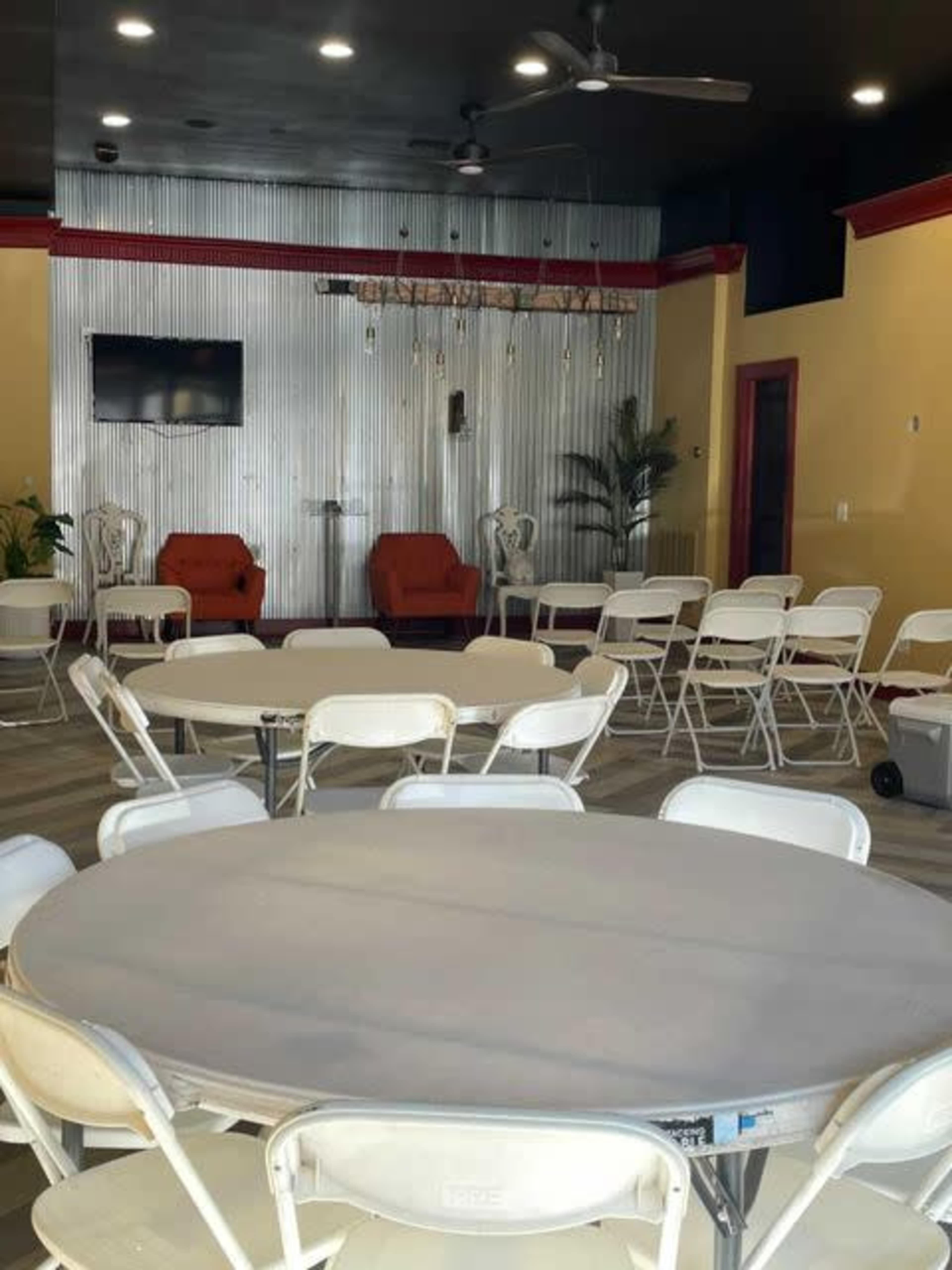 A spacious indoor venue with round tables and white folding chairs arranged for an event, featuring a television and decorative lighting in the background.