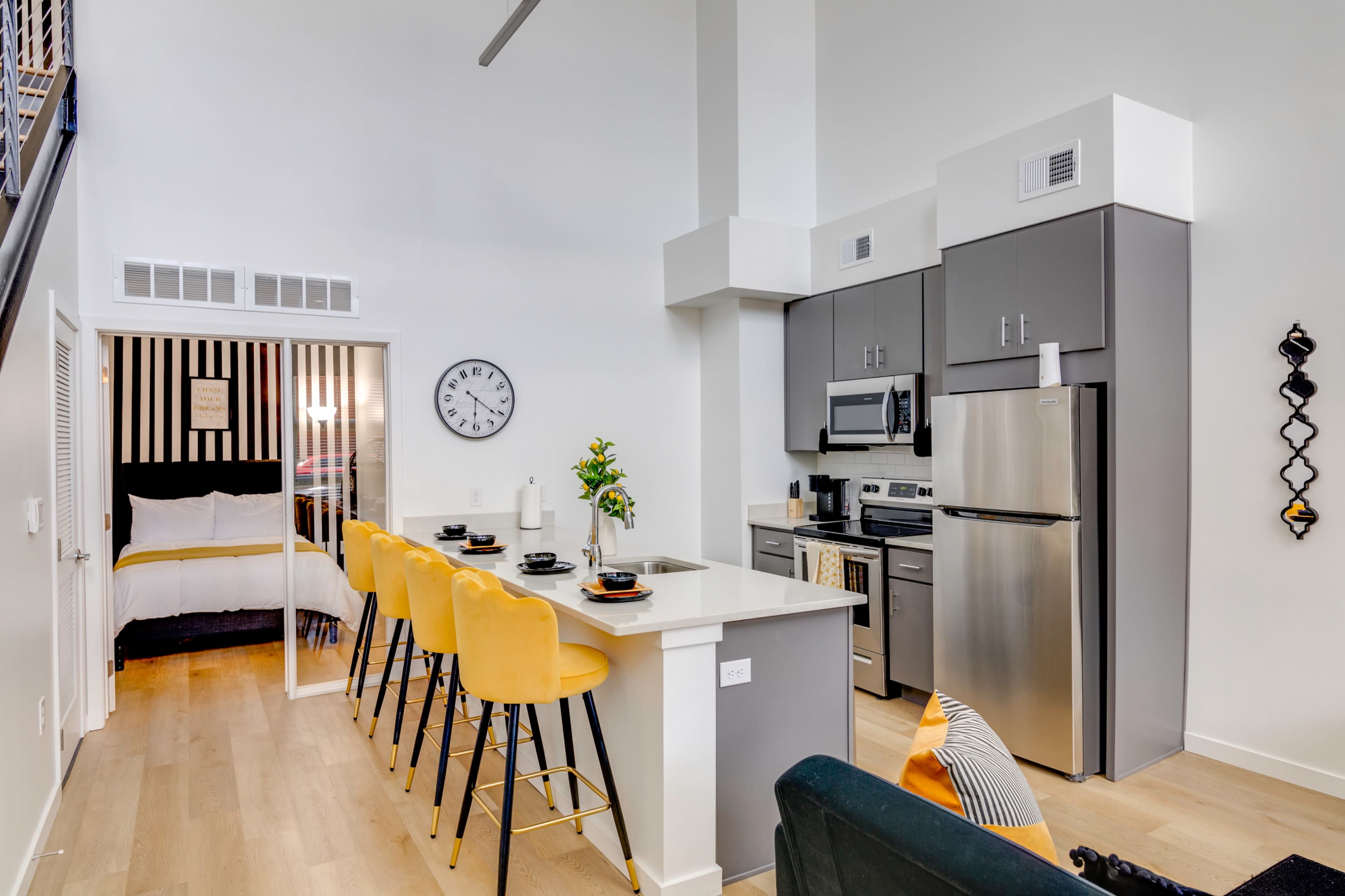 A modern kitchen area with stainless steel appliances, a breakfast bar with yellow stools, and a view into a bedroom with a black and white striped feature wall.