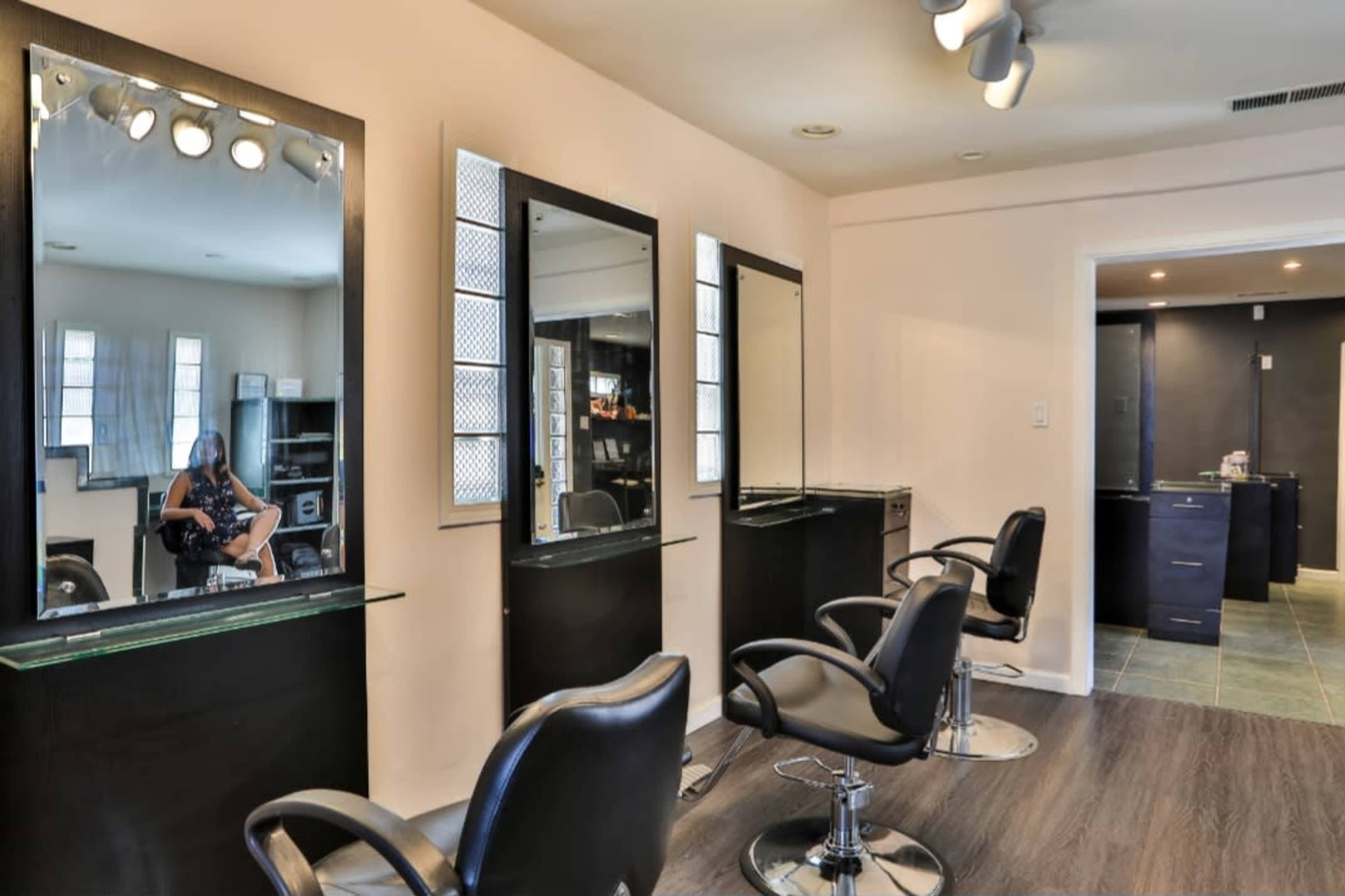A modern hair salon with three styling stations, each featuring a mirror and a black salon chair, and a customer seated in one of the mirrors.