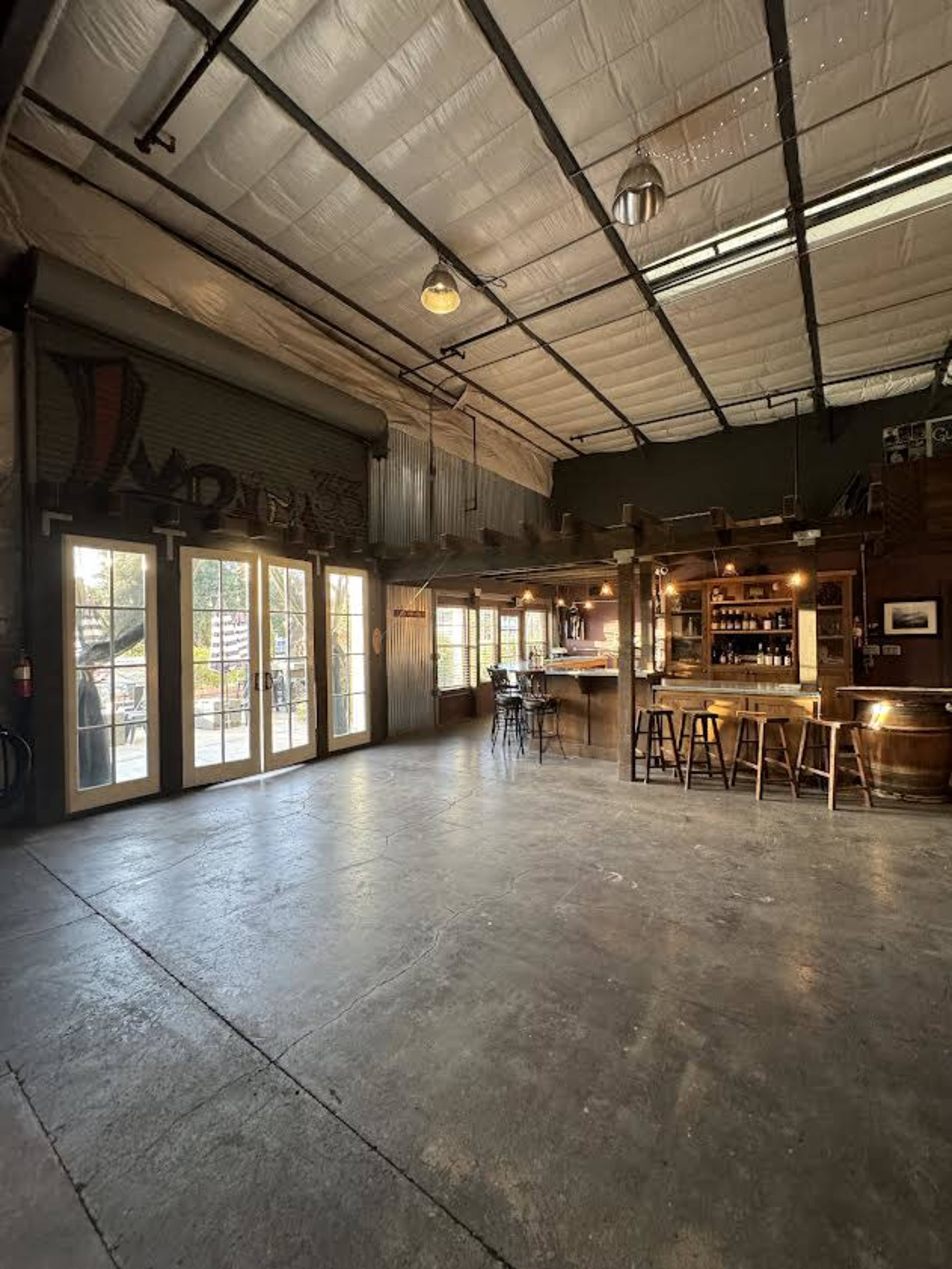 The interior of a spacious bar area features a wooden counter with seating, large glass doors leading outside, and exposed beams under a high ceiling.