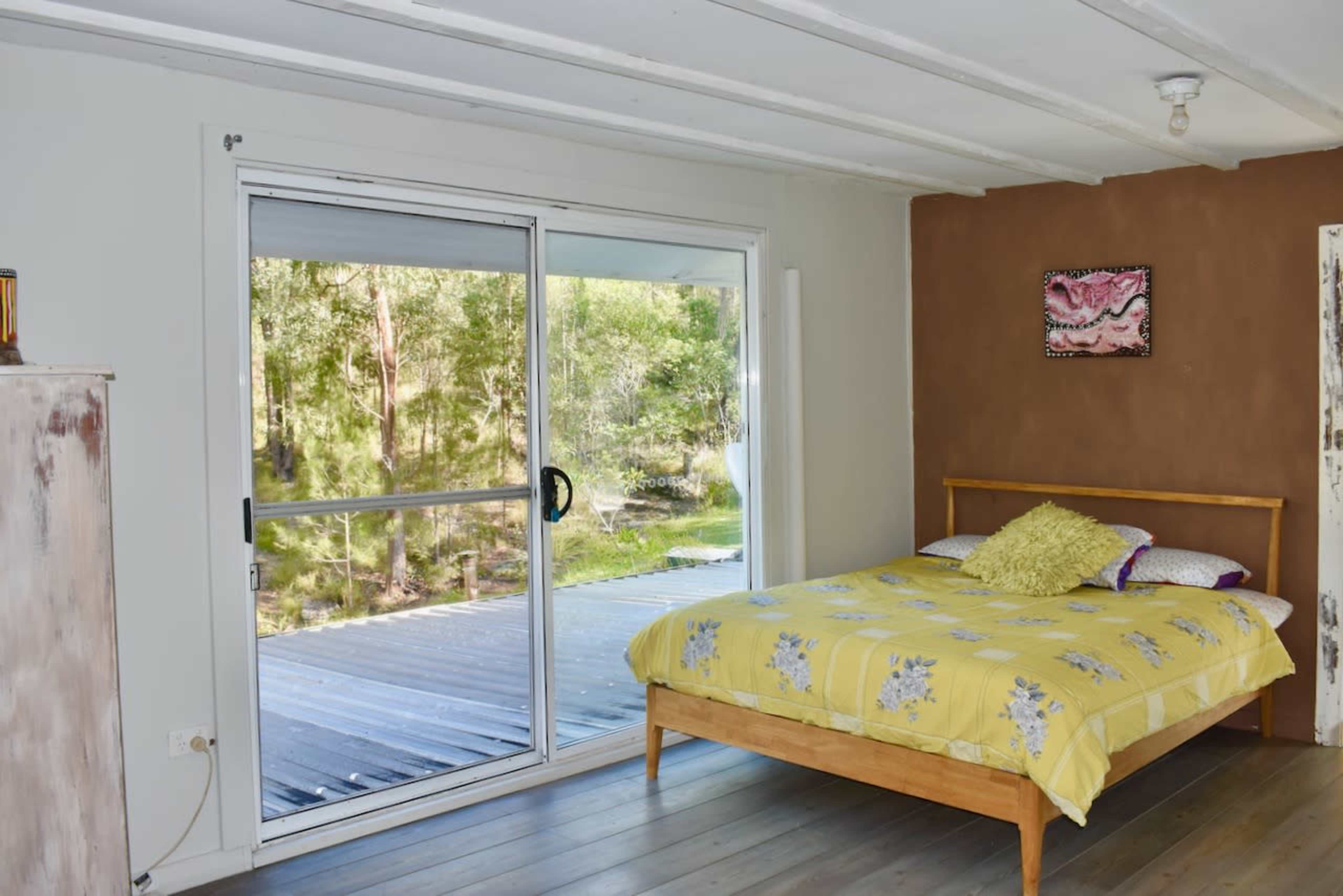 The image shows a bedroom with a wooden bed featuring a yellow floral bedspread, a sliding glass door leading to an outdoor deck, and a wall painted in a brown hue.
