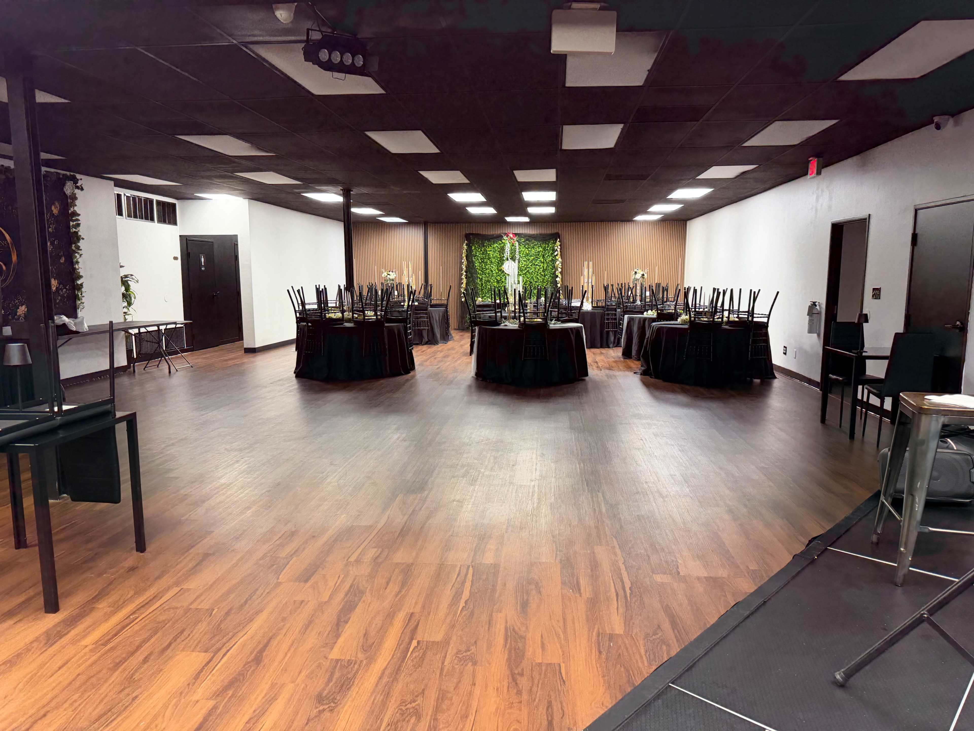 The image shows a spacious event hall with multiple round tables covered in black tablecloths, surrounded by black chairs, and decorated with greenery on one wall.