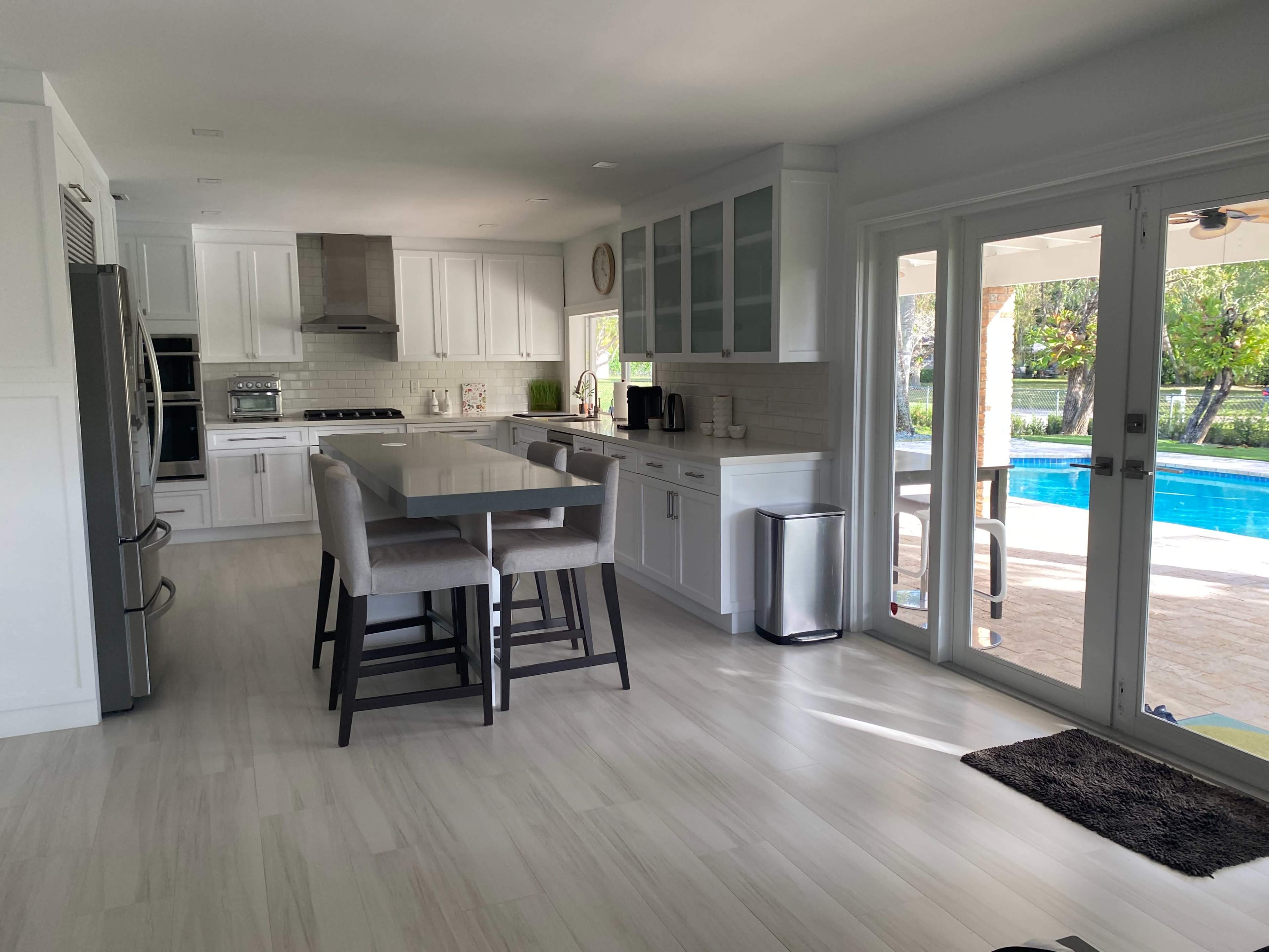 A modern kitchen with white cabinetry, a large central island with four barstools, and sliding glass doors leading to an outdoor pool area.